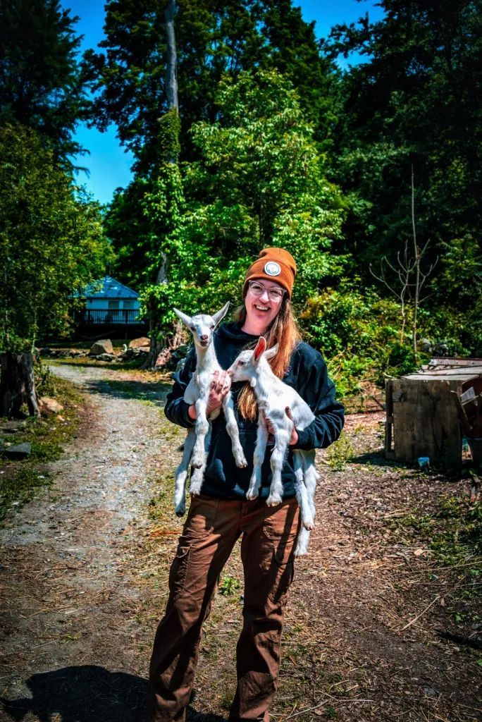 Woman holding two baby goats, smiling, wearing a hat and brown pants, outdoors.