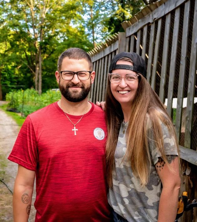 A man and woman standing together outdoors. The man wears glasses and a red shirt. The woman wears a camo shirt and a cap.