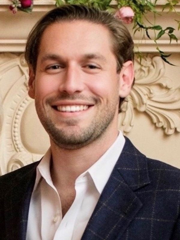 A man in a suit and white shirt is smiling in front of a fireplace.