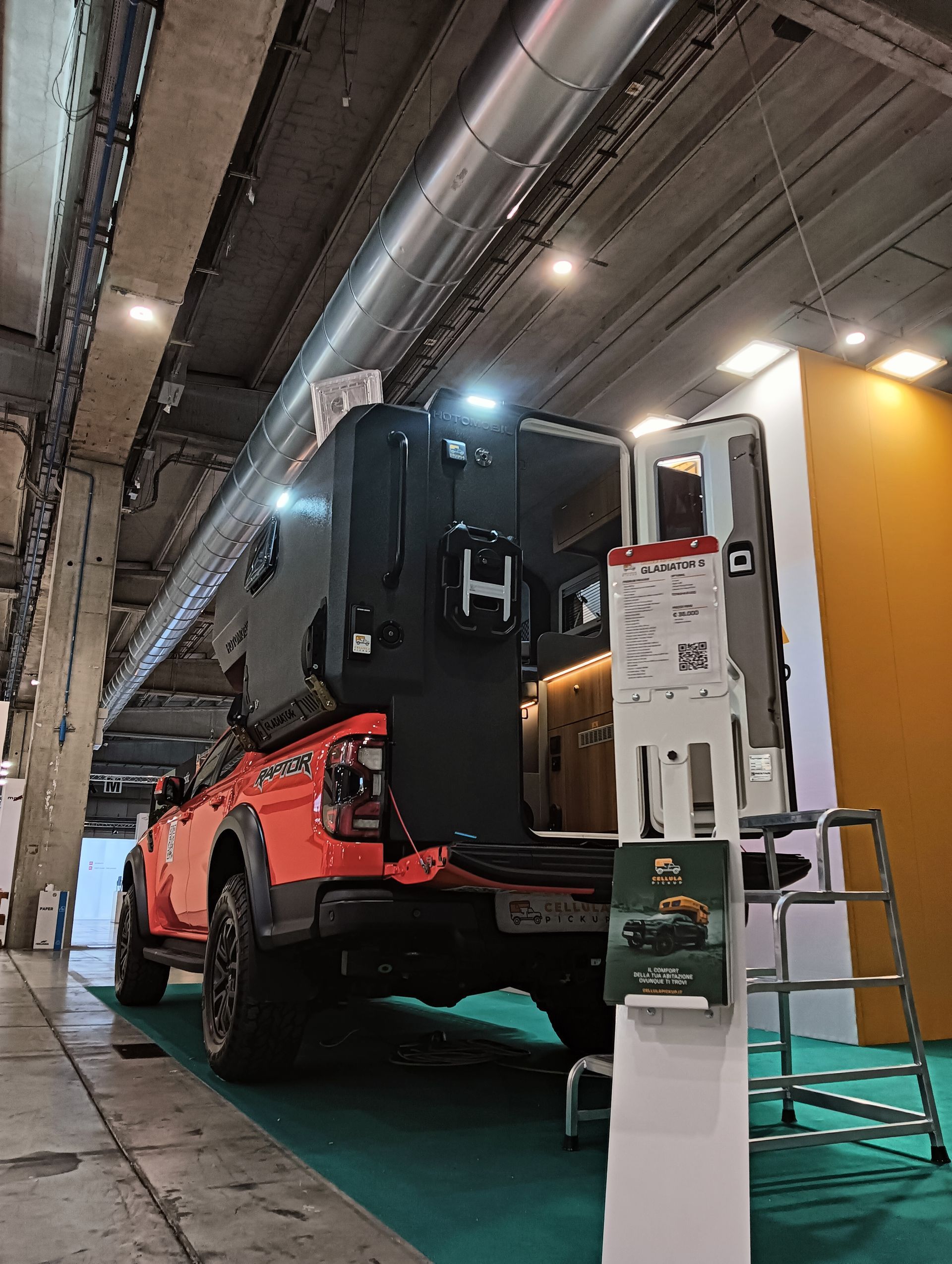 Un pick-up arancione con una scocca da camper nera in una sala espositiva al coperto.