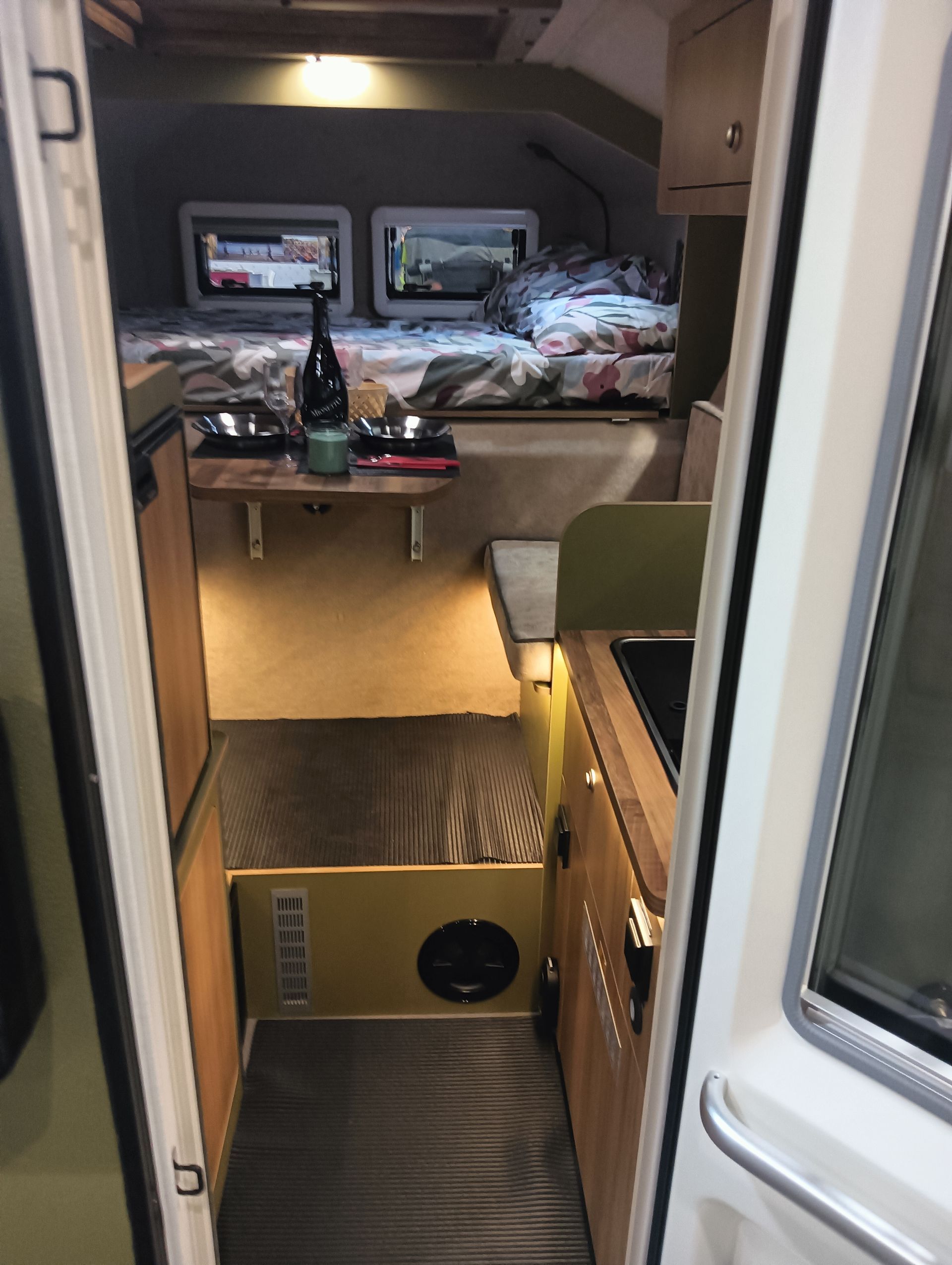 Interno di un camper con letto, cucina e tavolino. Colori neutri con dettagli in legno.