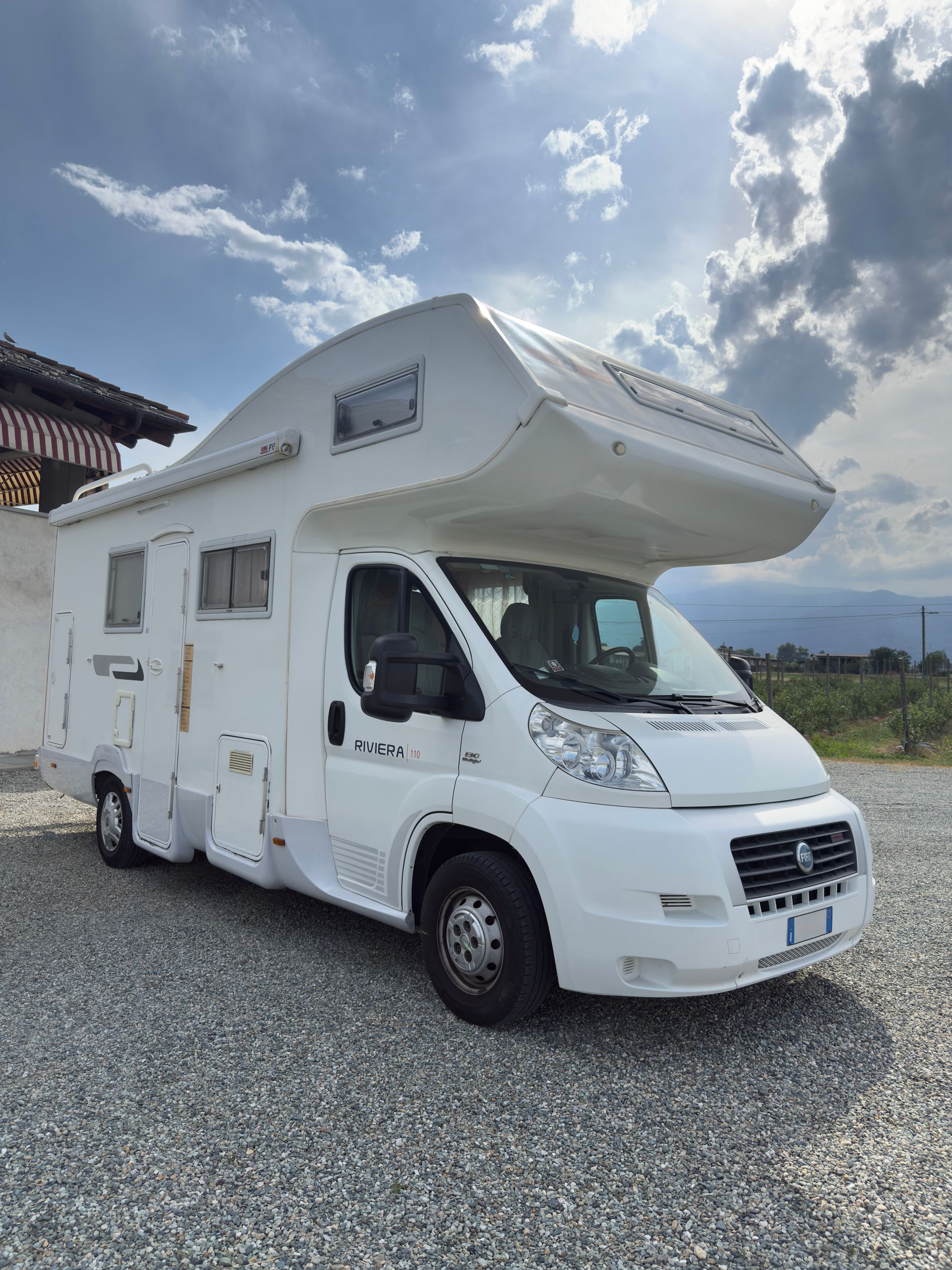 Camper bianco parcheggiato sulla ghiaia, giornata di sole, cielo azzurro con nuvole.
