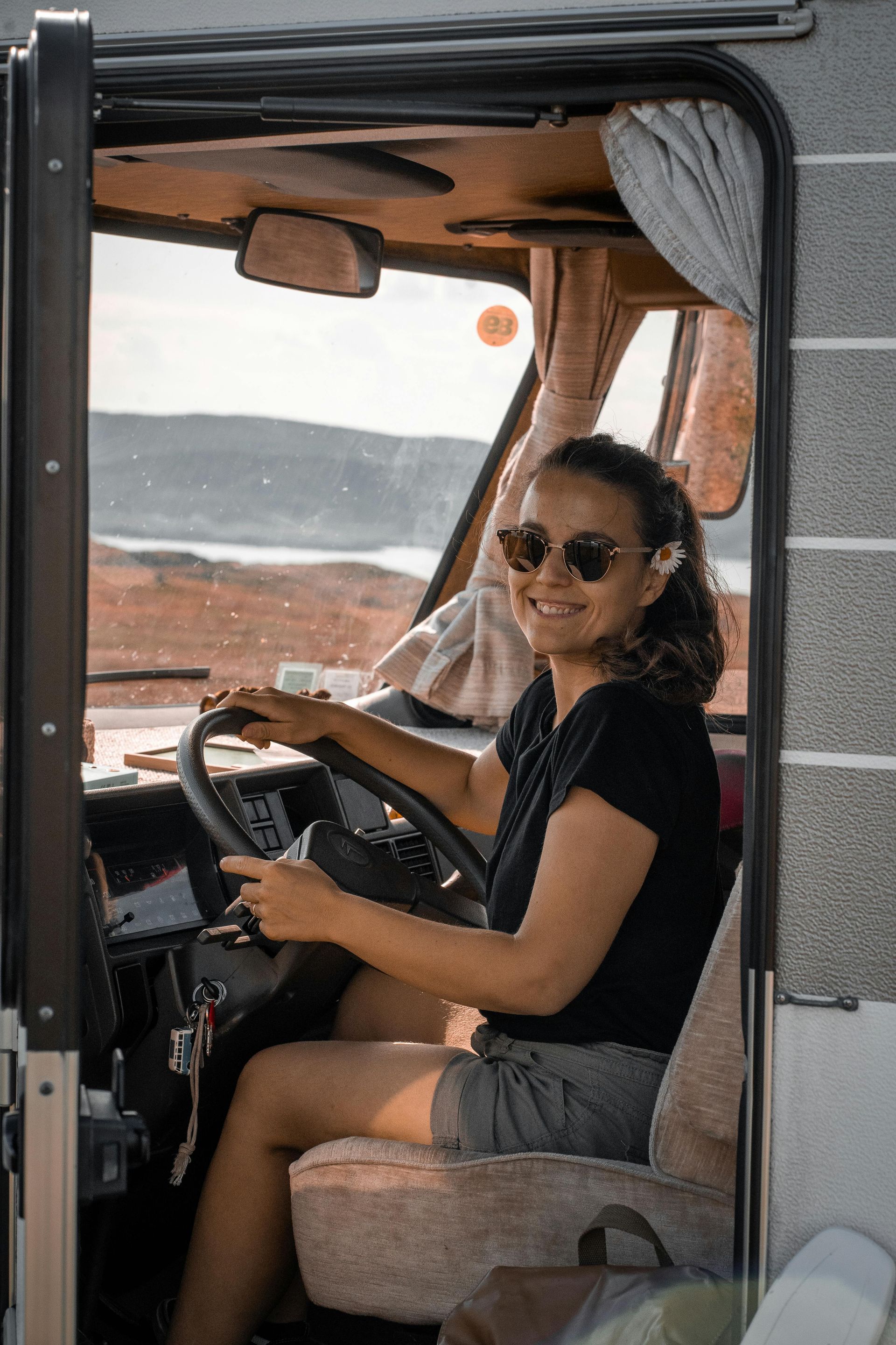 Una donna sorride mentre guida un camper. Indossa occhiali da sole e pantaloncini, ed è seduta al volante.