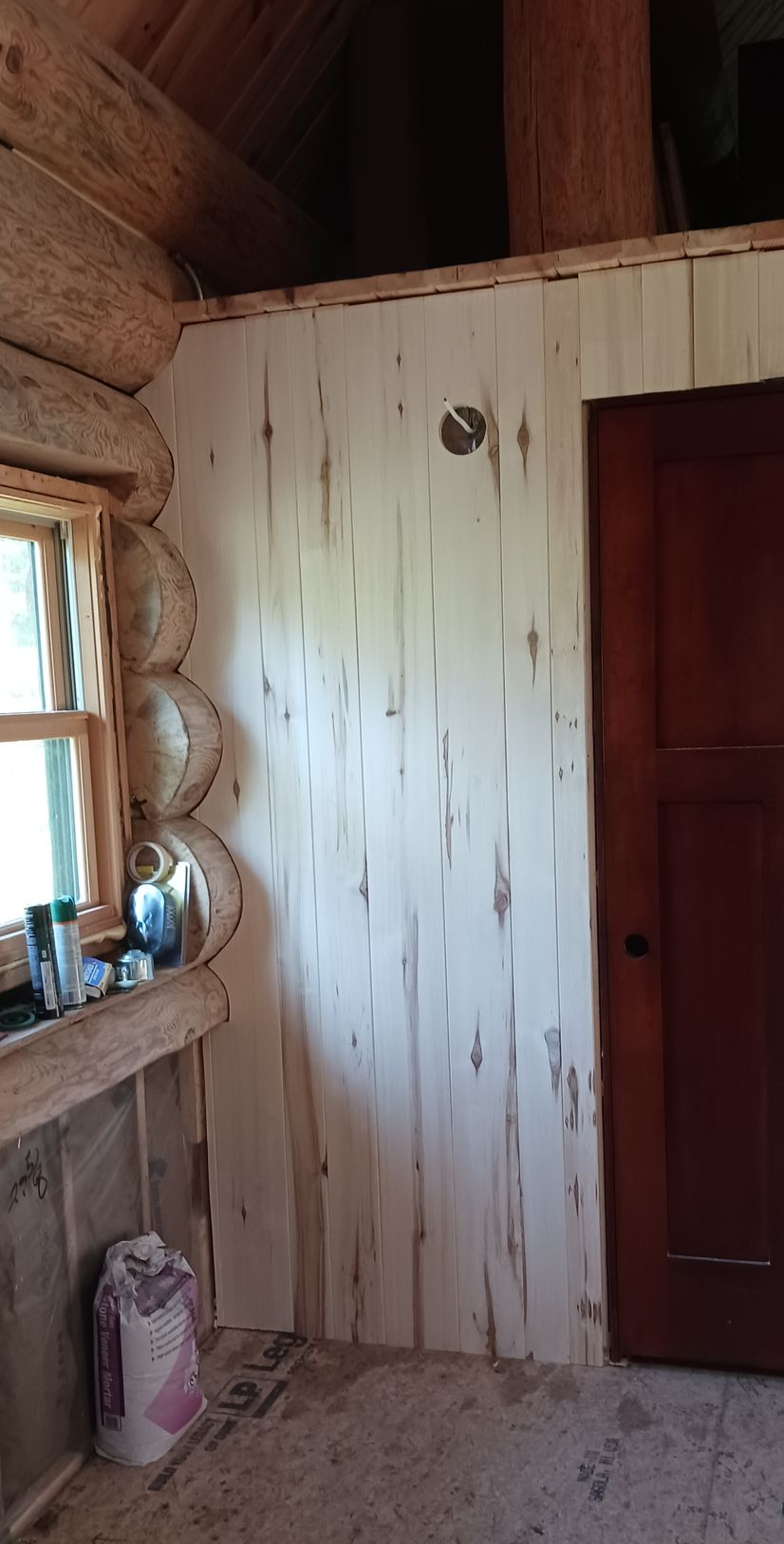 A whitewashed wooden wall in a rustic log cabin, featuring a window on the left, a dark wooden door, and exposed logs above.
