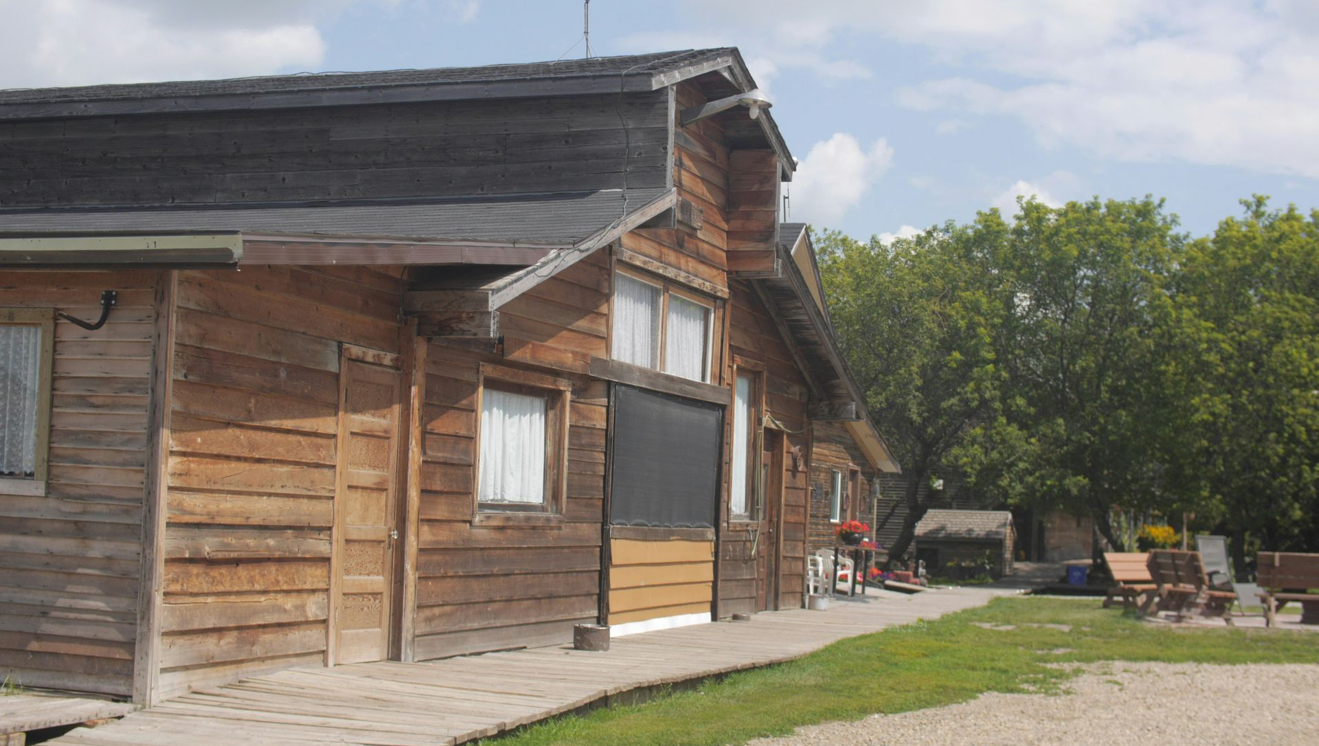 Wooden cabin with a gravel courtyard, green lawn, and trees under a cloudy sky