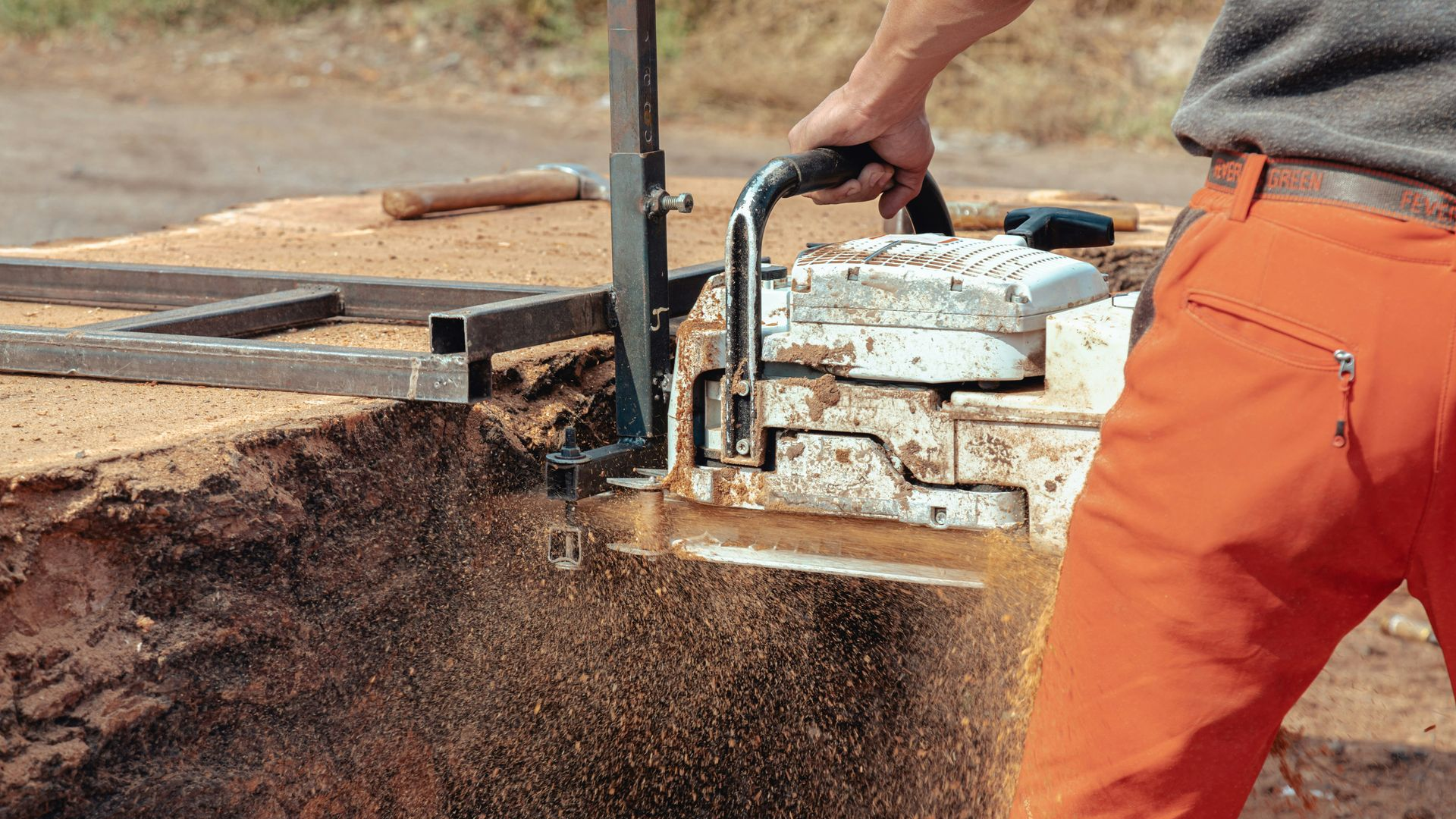 Worker in orange pants using a chainsaw to cut a log outdoors, sawdust flying