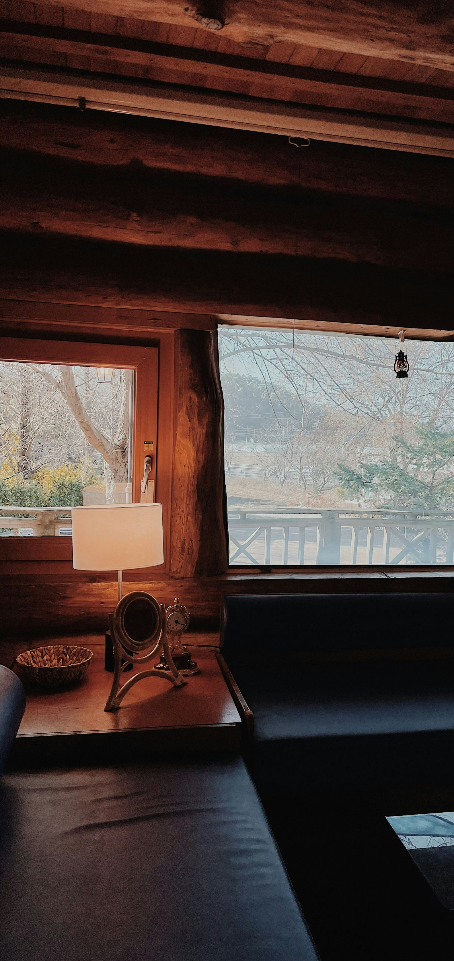 Cozy cabin interior with wooden beams, a lamp on a table, and a large window overlooking snowy trees and a lake