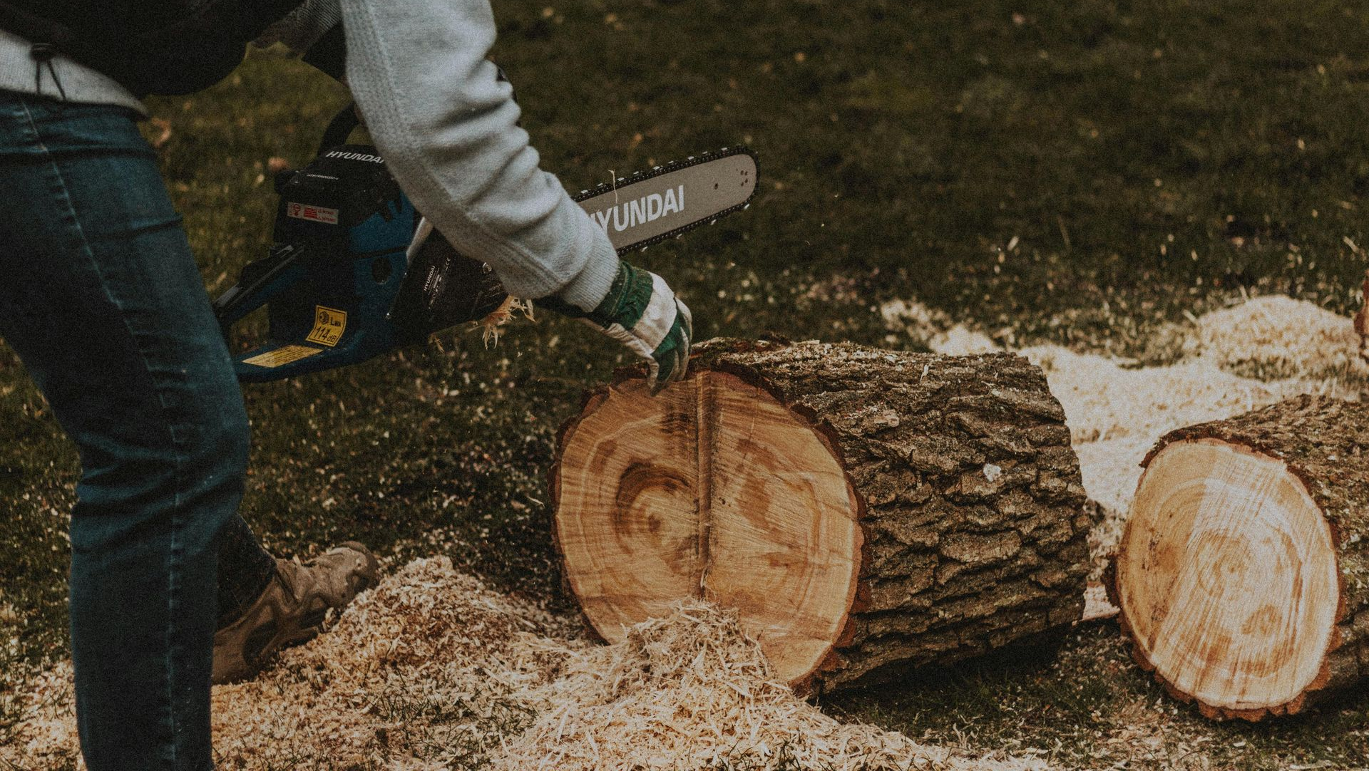 Person in jeans and gloves using a chainsaw to cut a log outdoors.
