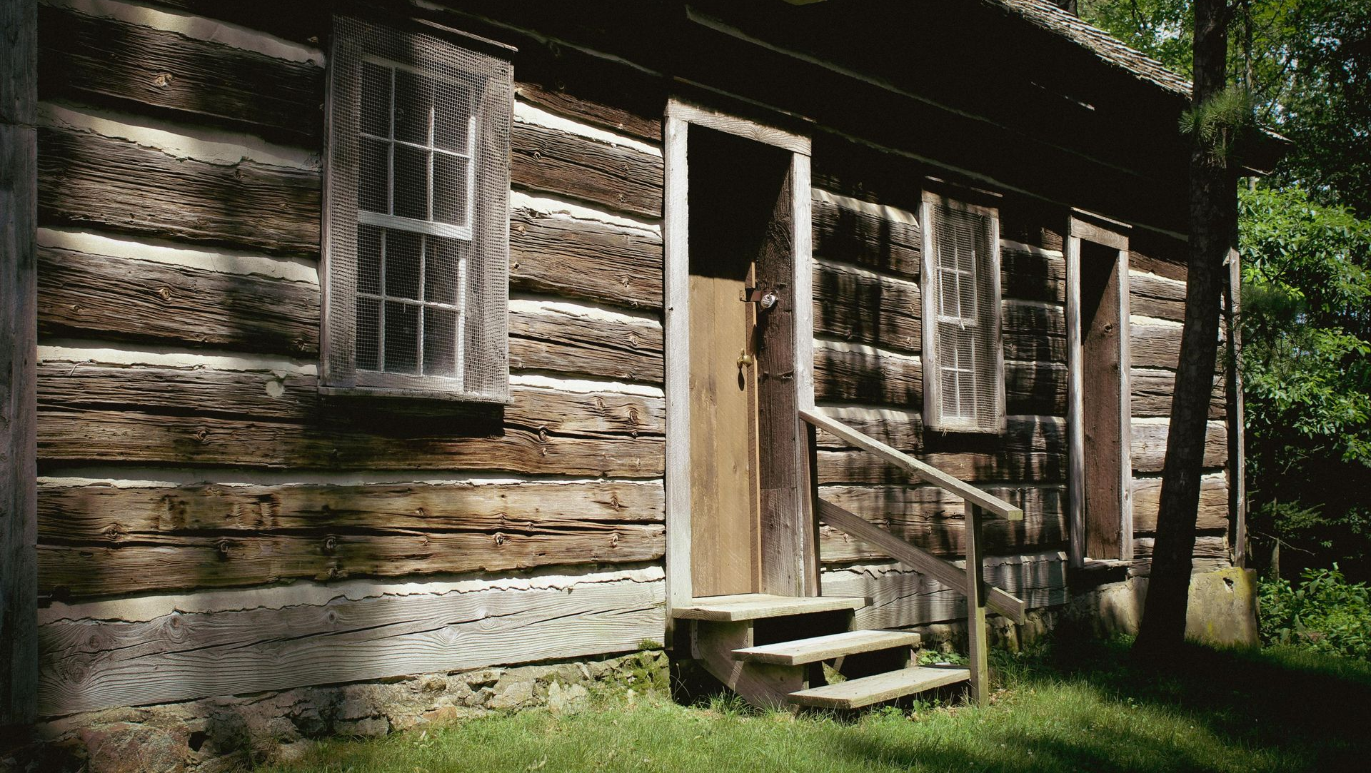 Rustic log cabin with open door, small porch, and steps in a wooded setting