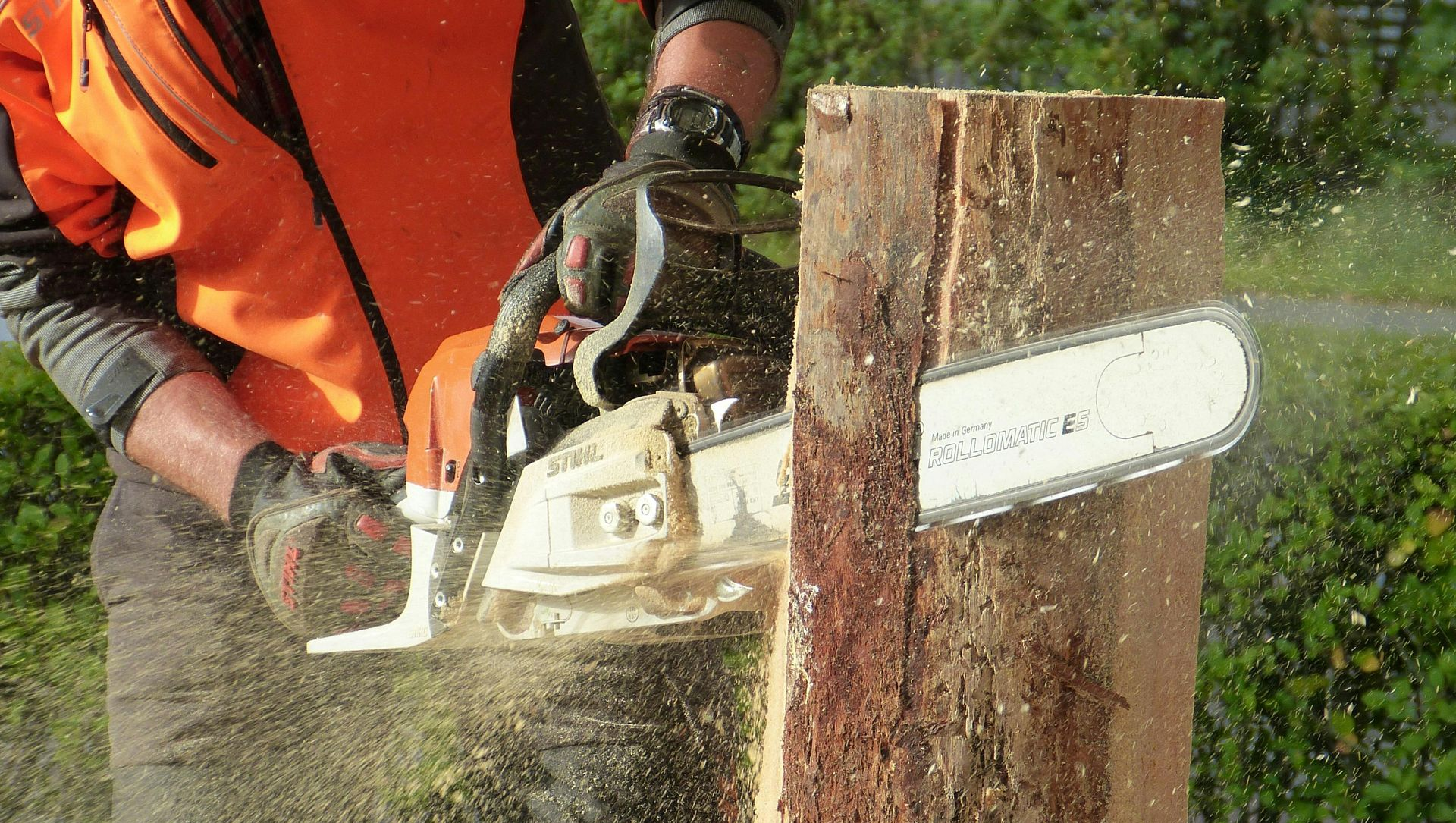 Person using a chainsaw to cut through a wooden post, with sawdust flying outdoors