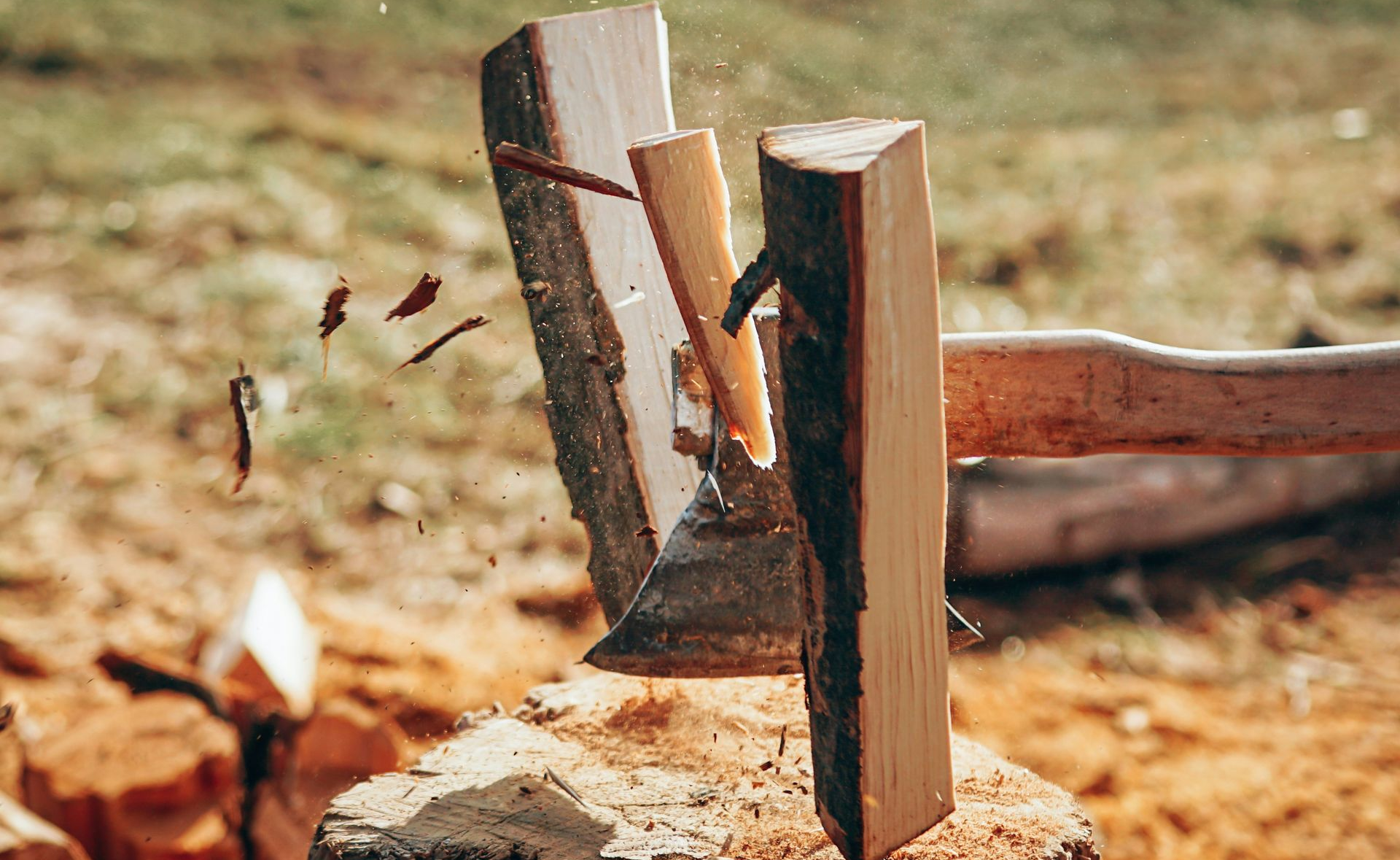 A hatchet splits a log on a stump outdoors, with wood chips flying.