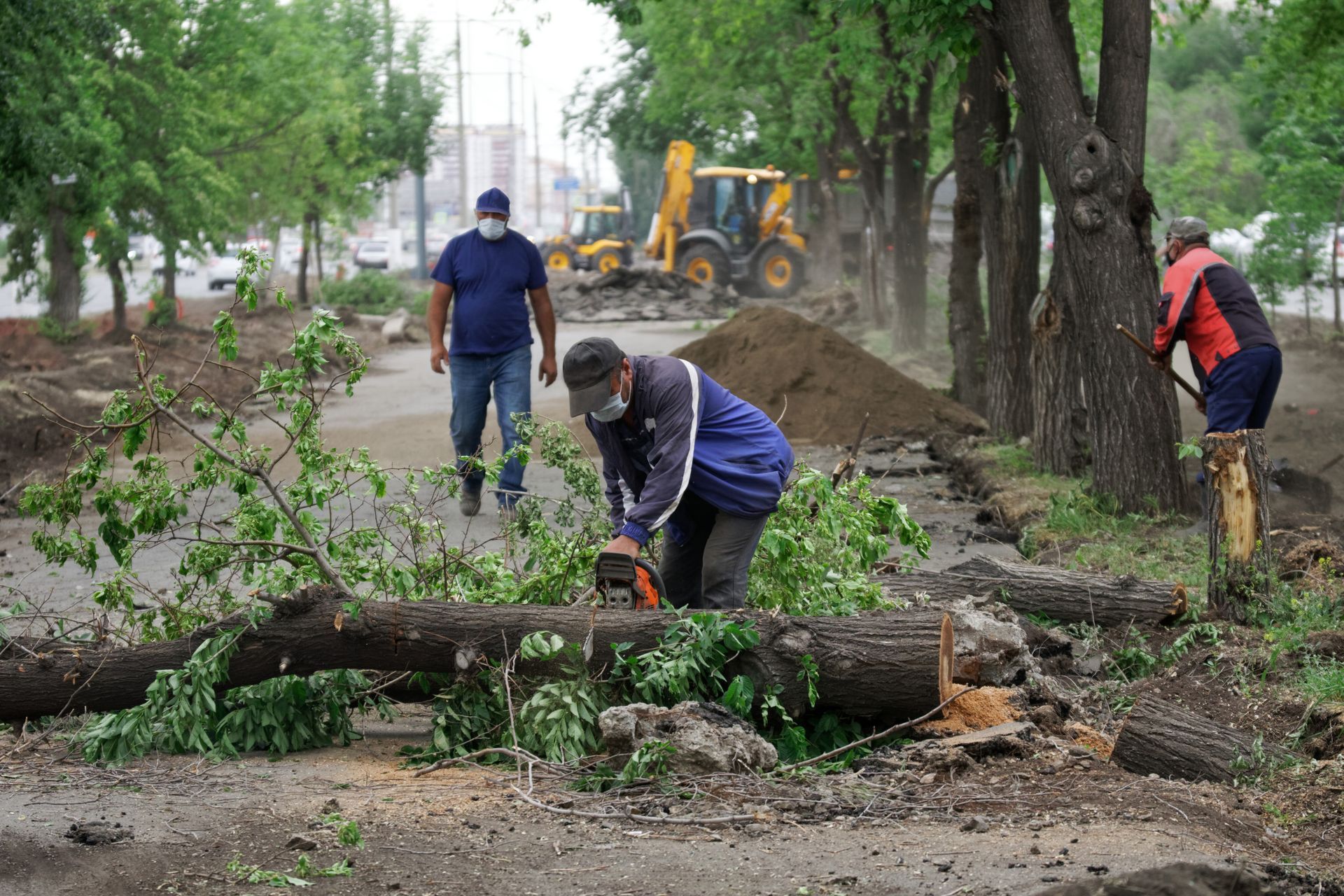 Workers clearing fallen tree limbs along a muddy roadside with a backhoe in the background Workers clearing fallen tree limbs along a muddy roadside with a backhoe in the background