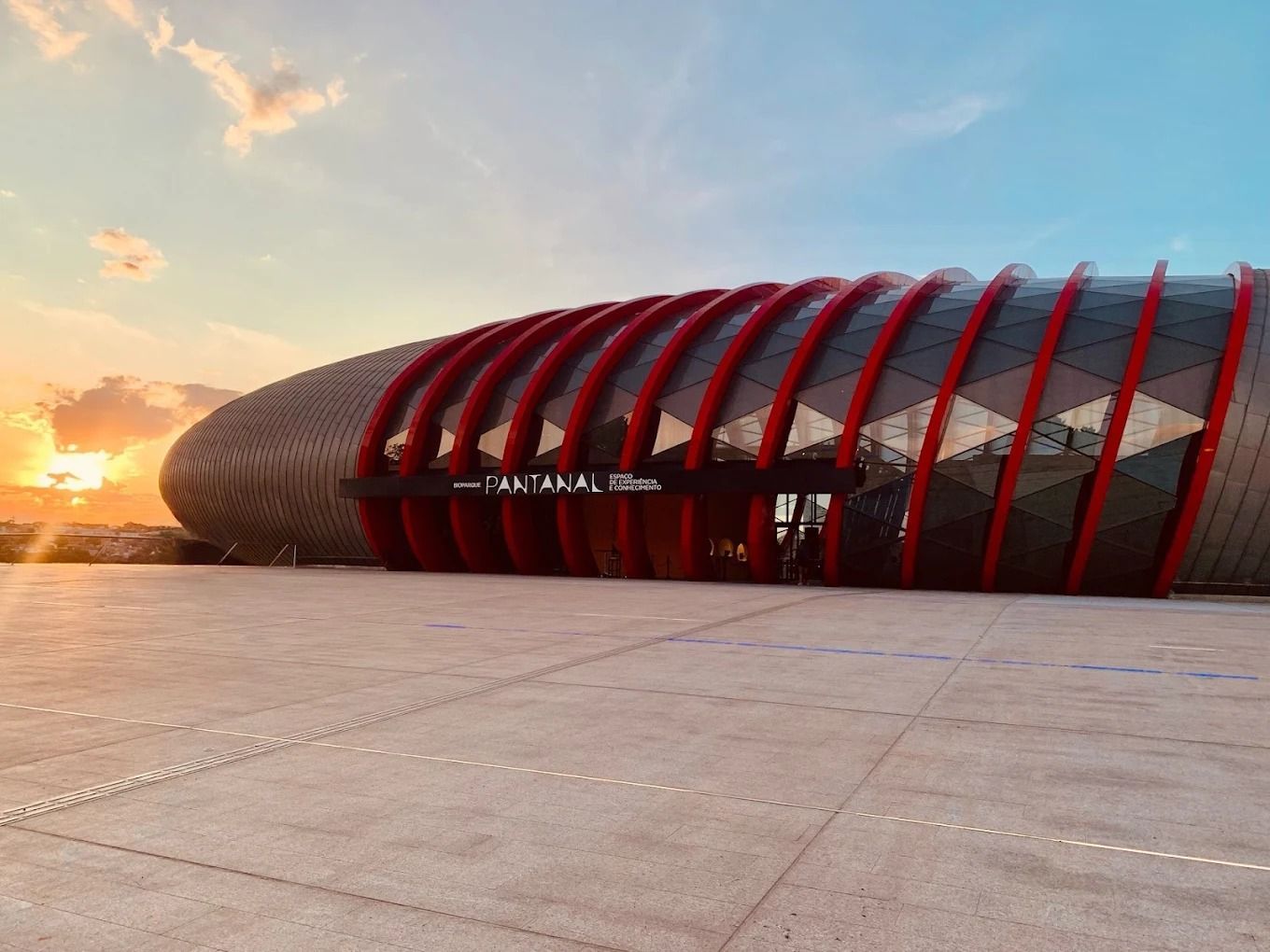 Prédio moderno com detalhes em vermelho e painéis de vidro sob o pôr do sol. O texto diz 