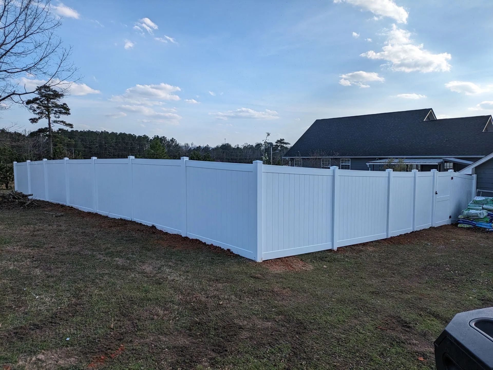 White vinyl fence encloses a grassy backyard on a sunny day. A house is visible in the background.