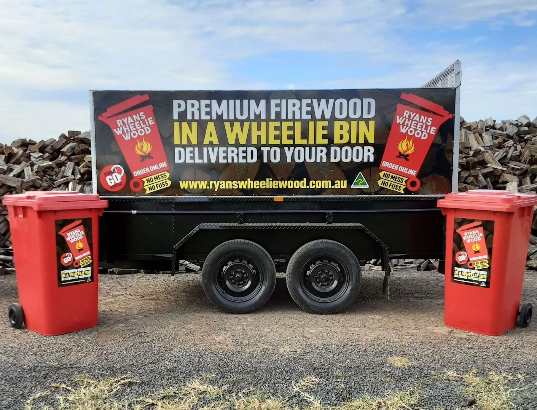 Trailer advertising premium firewood delivery, flanked by two red wheelie bins on a gravel lot