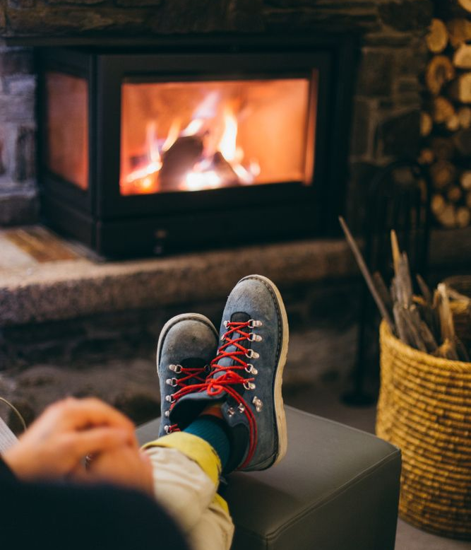 Feet in gray sneakers with red laces resting by a lit fireplace in a cozy living room.