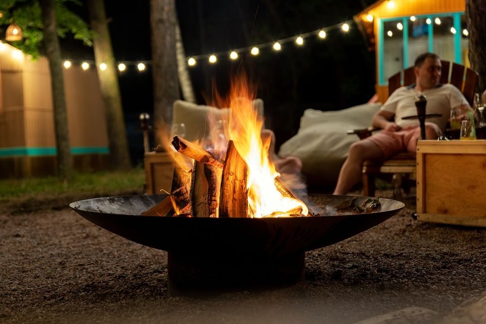 Campfire glowing in a backyard at night, with string lights and a person relaxing on a chair nearby.