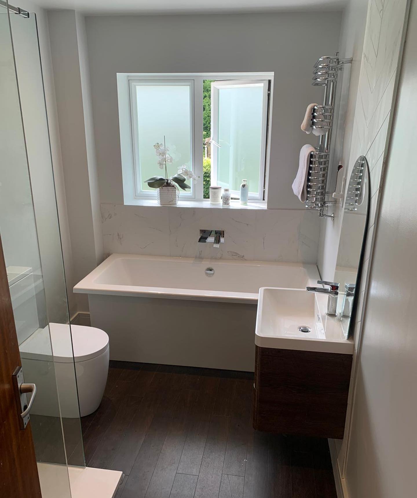 Modern bathroom with a bathtub, toilet, sink, and window with frosted glass. Dark wooden floor.