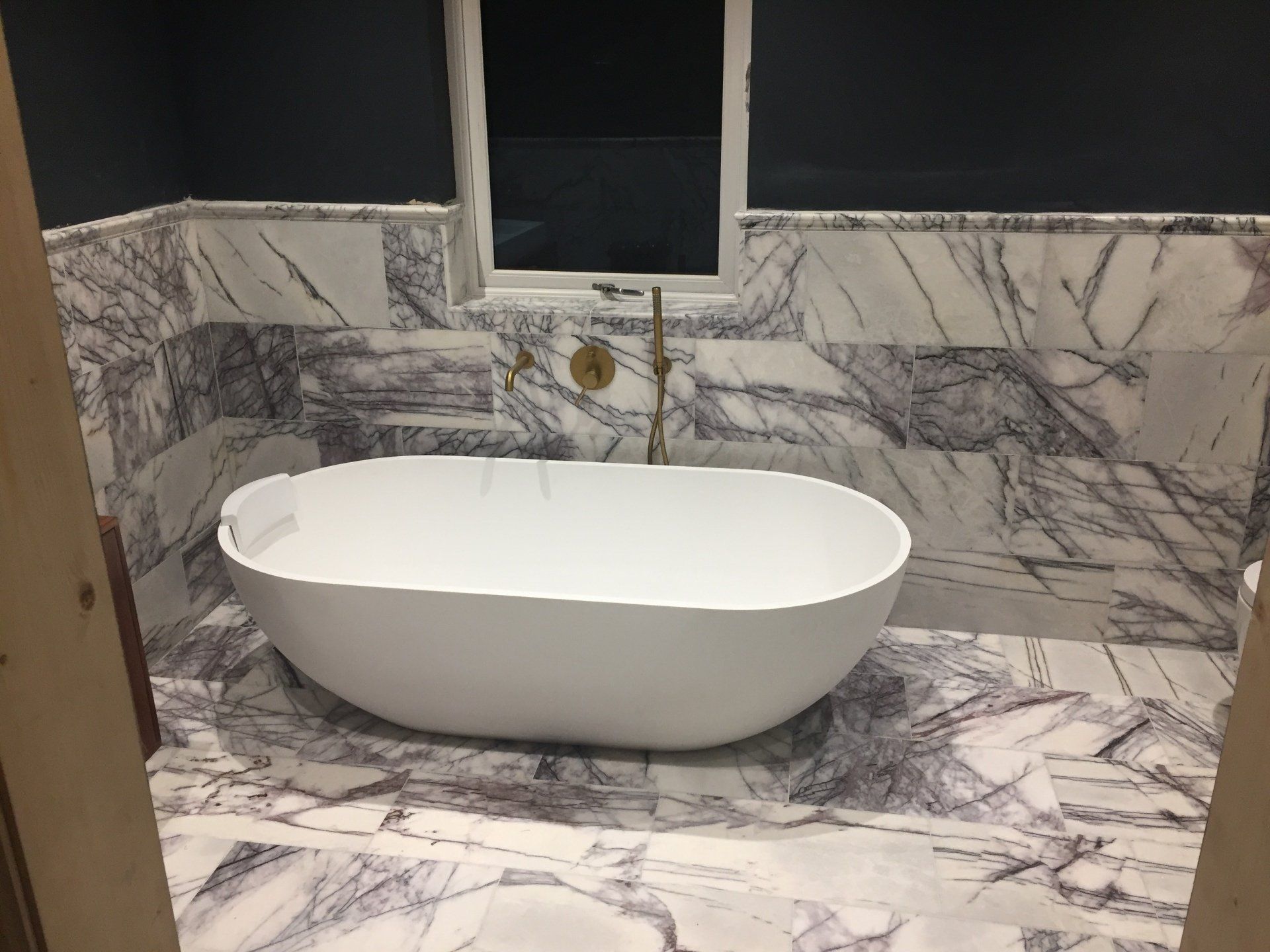 Oval bathtub surrounded by marble tile in a bathroom. Gold faucet and dark blue wall above.