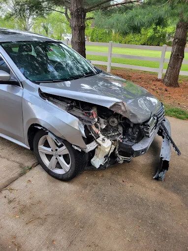 Silver car with front-end collision damage parked by a white fence and trees.