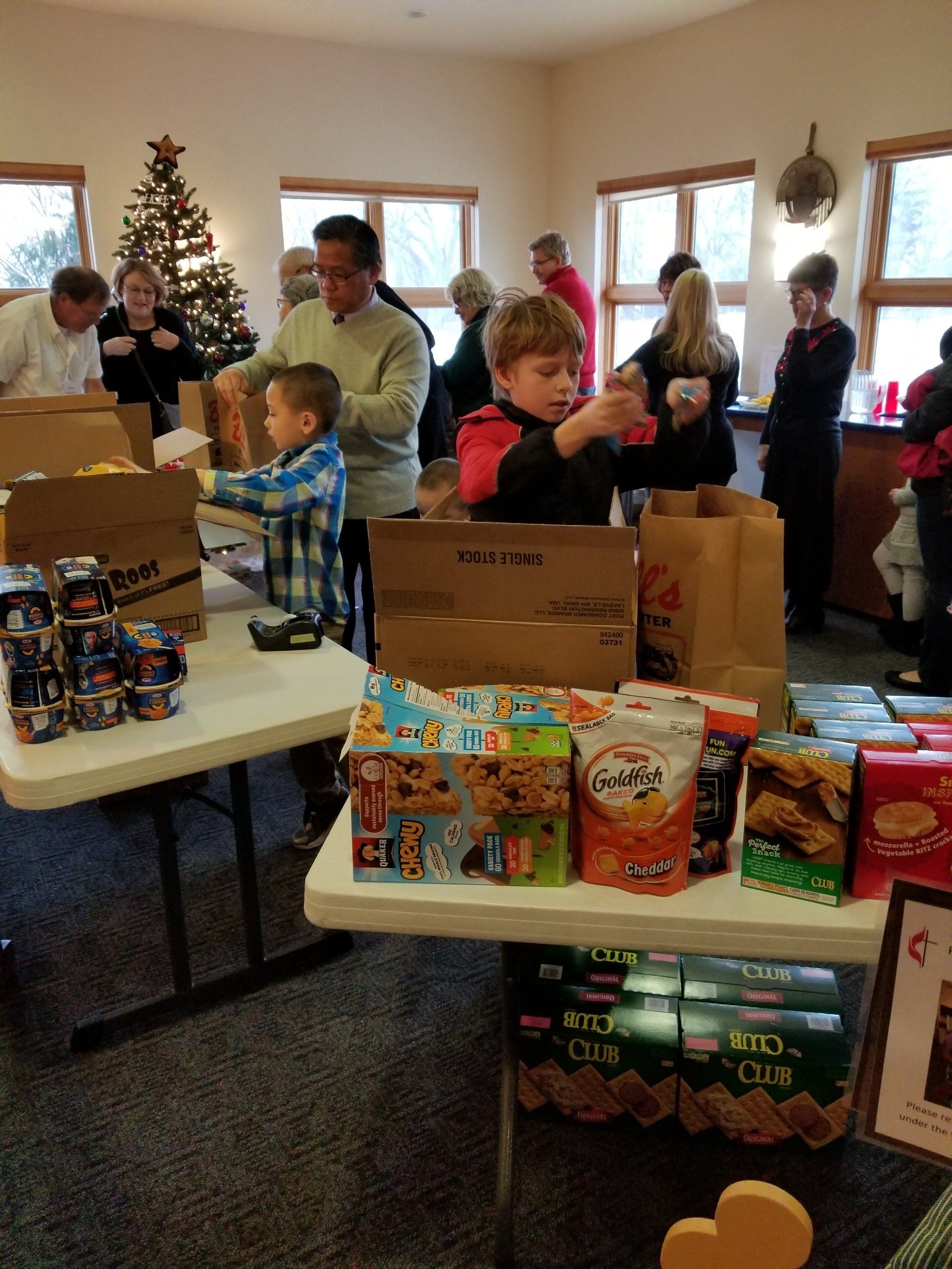 People packing food items at tables for donation, near a Christmas tree.