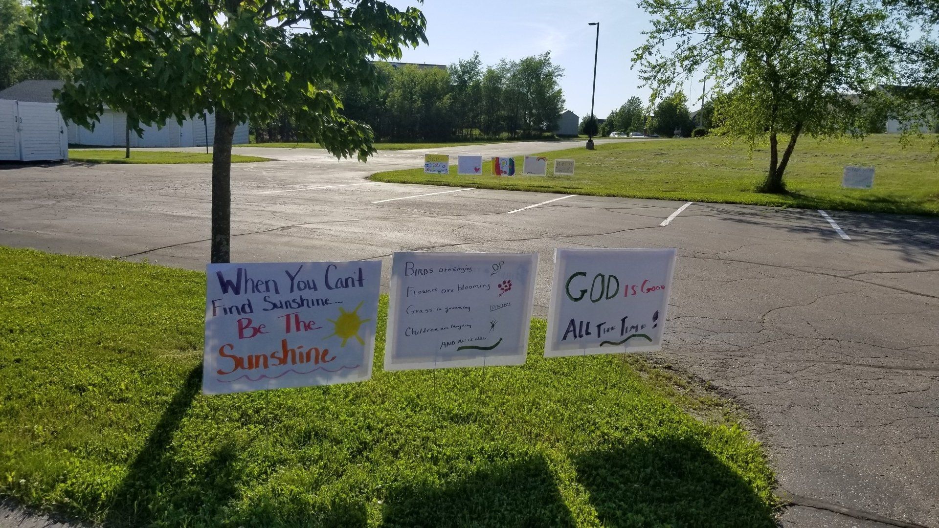 Hand-written signs with messages in a grassy parking lot on a sunny day.