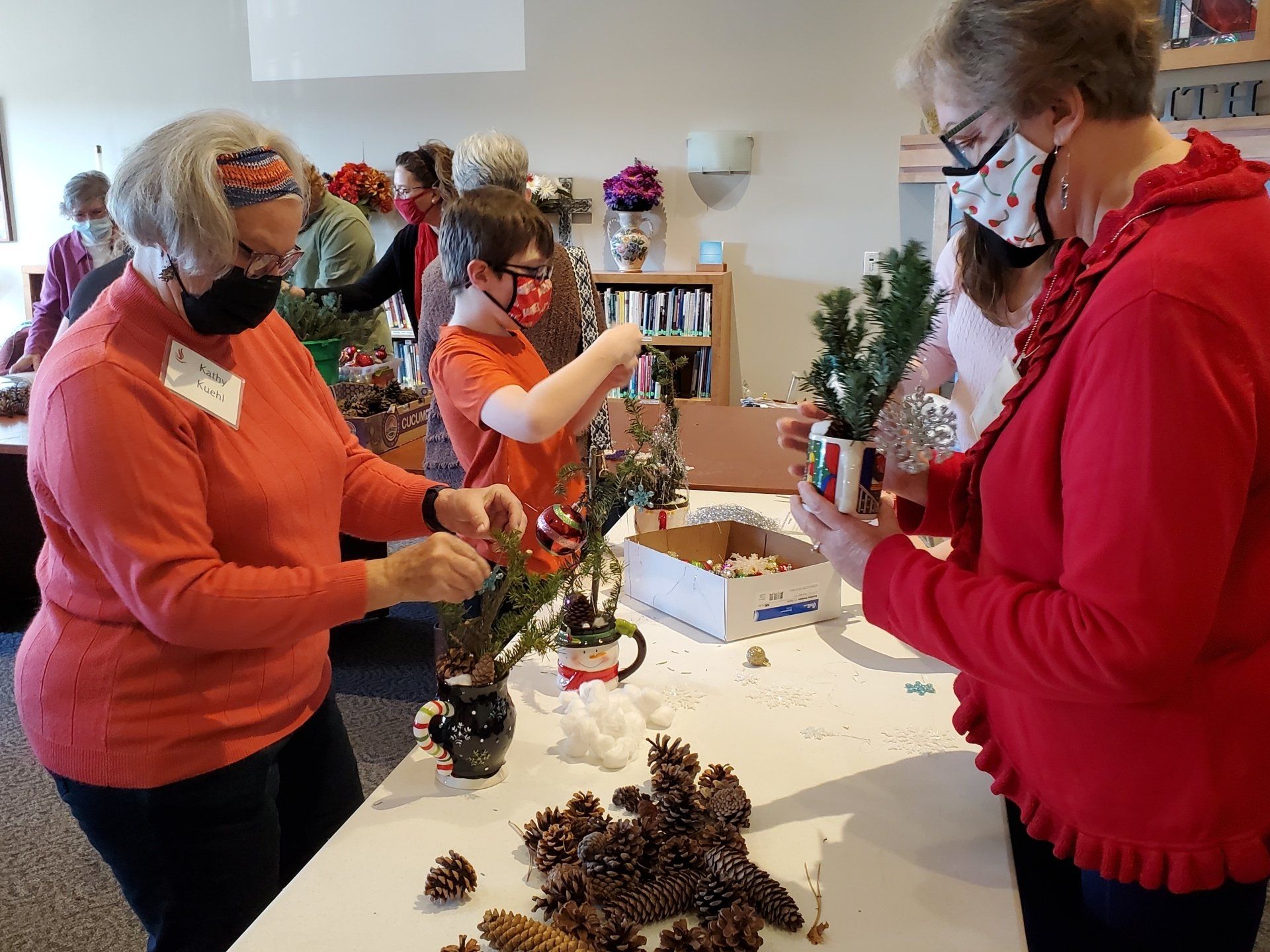 People crafting floral arrangements at a table. Several wear masks. Pine cones and flowers are visible.