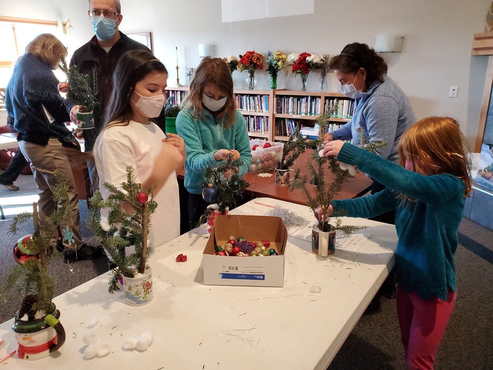 People, including children, decorating small Christmas trees at a table indoors.