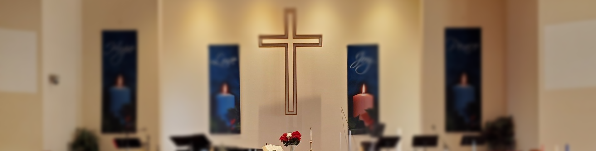 A wooden cross is centered in front of a church altar, with three blue banners and a flower arrangement below it.
