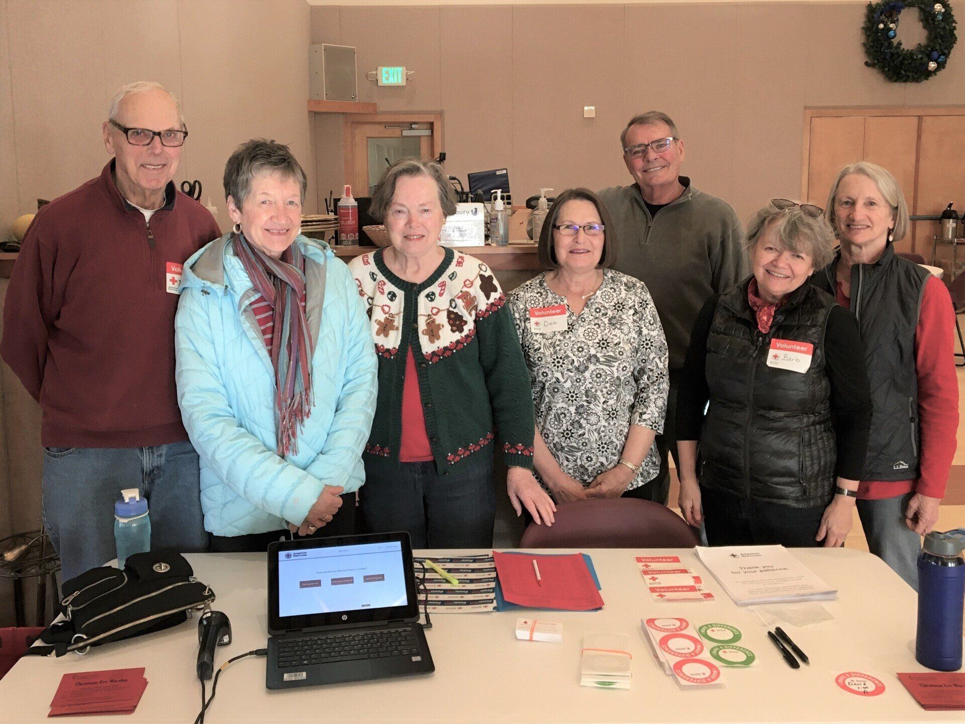 Group of people standing behind a table with a laptop, smiling. Indoor setting, Christmas decorations.
