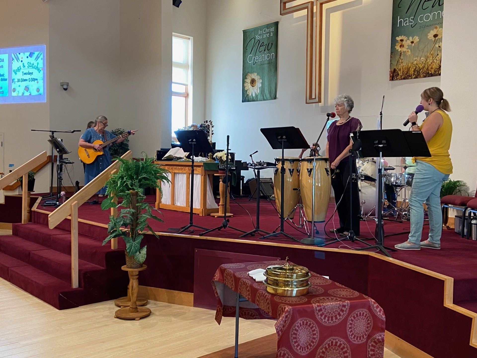 Band performing on a stage in a church. Musicians play guitar, congas, keyboard, and sing. A cross hangs on the back wall.