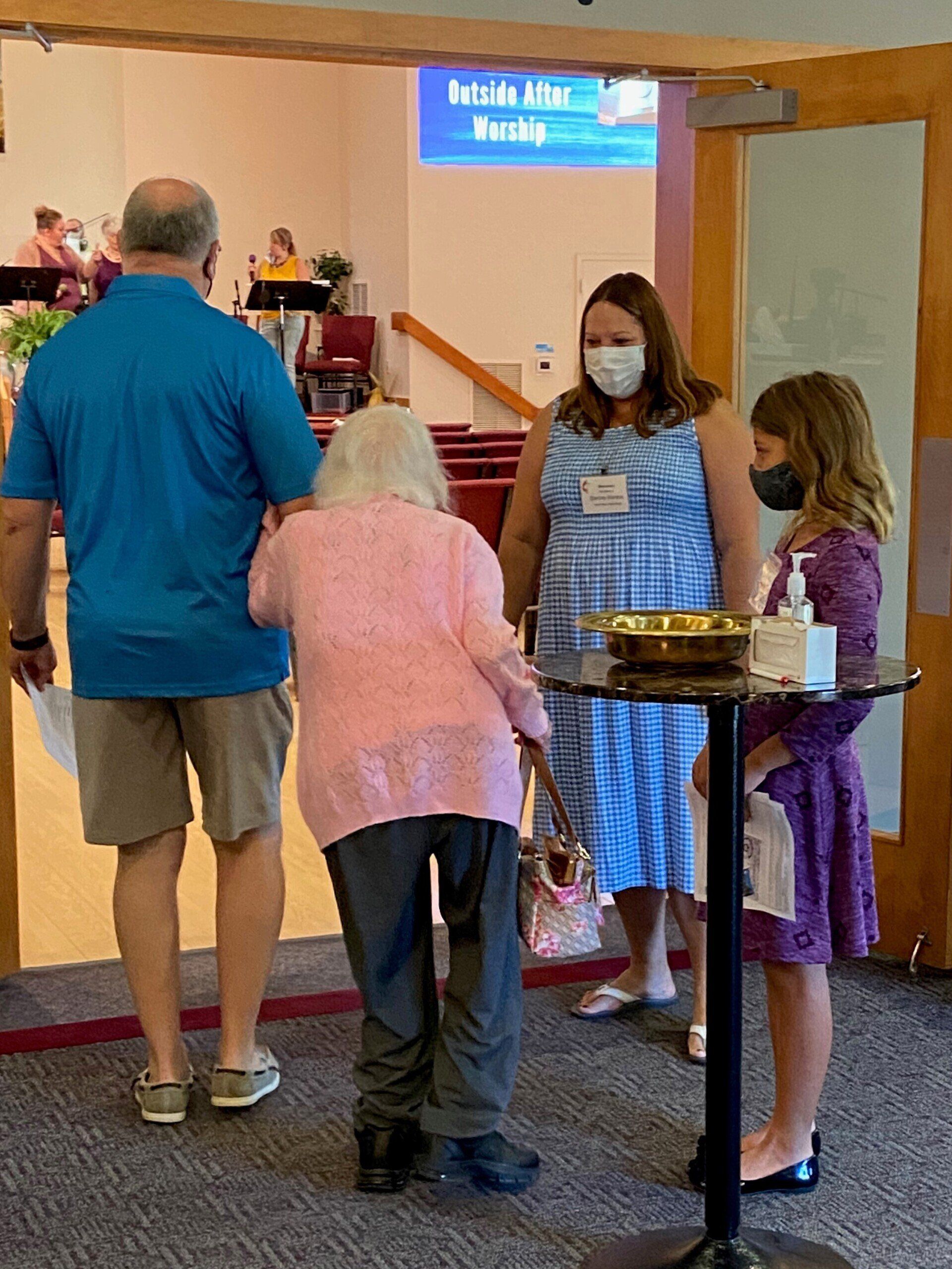 People entering a building, one using a cane, greeted by two others near a small table with sanitizer.
