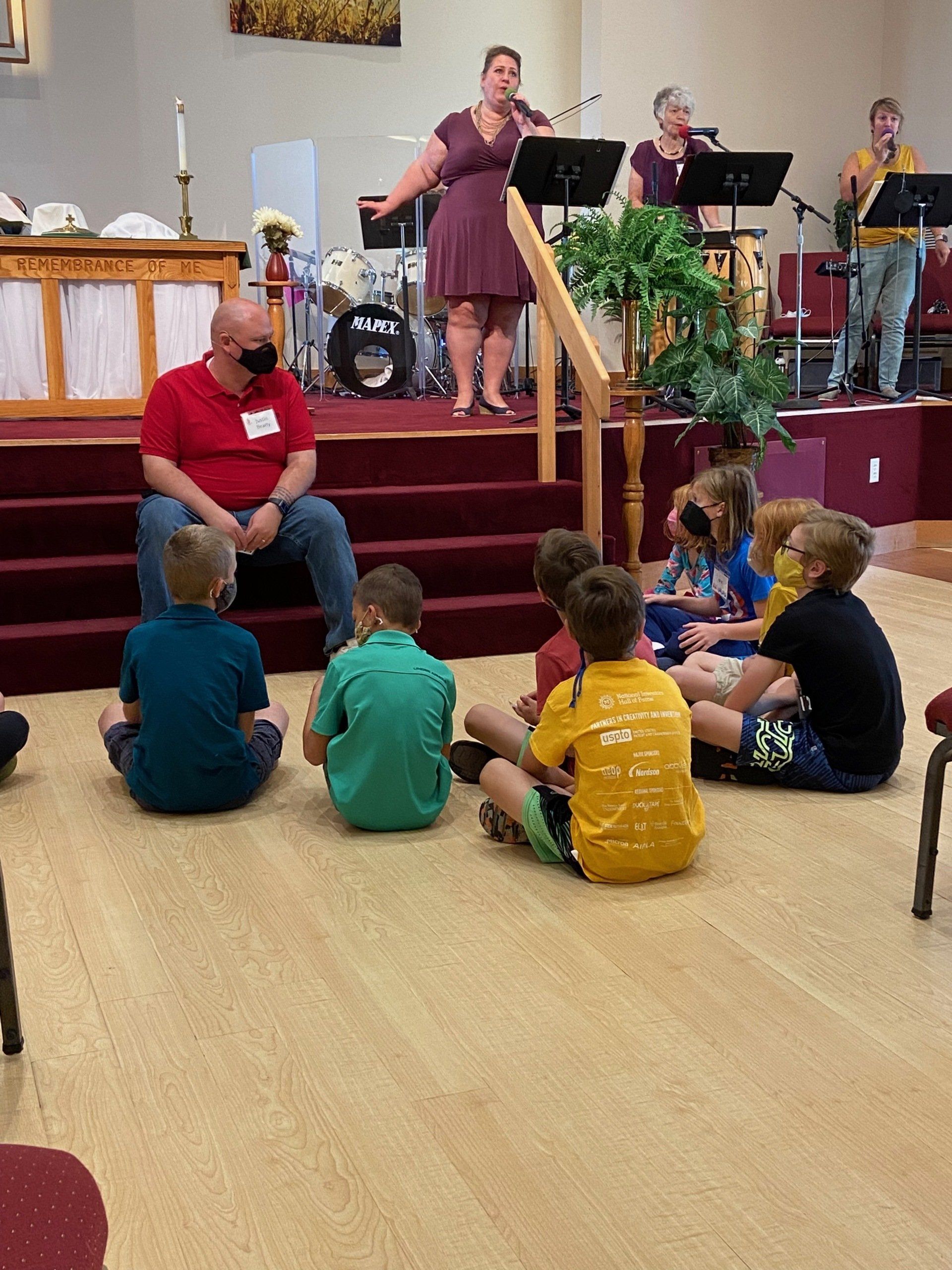 A group of children sits on the floor listening to a man and a band on a stage in a church.