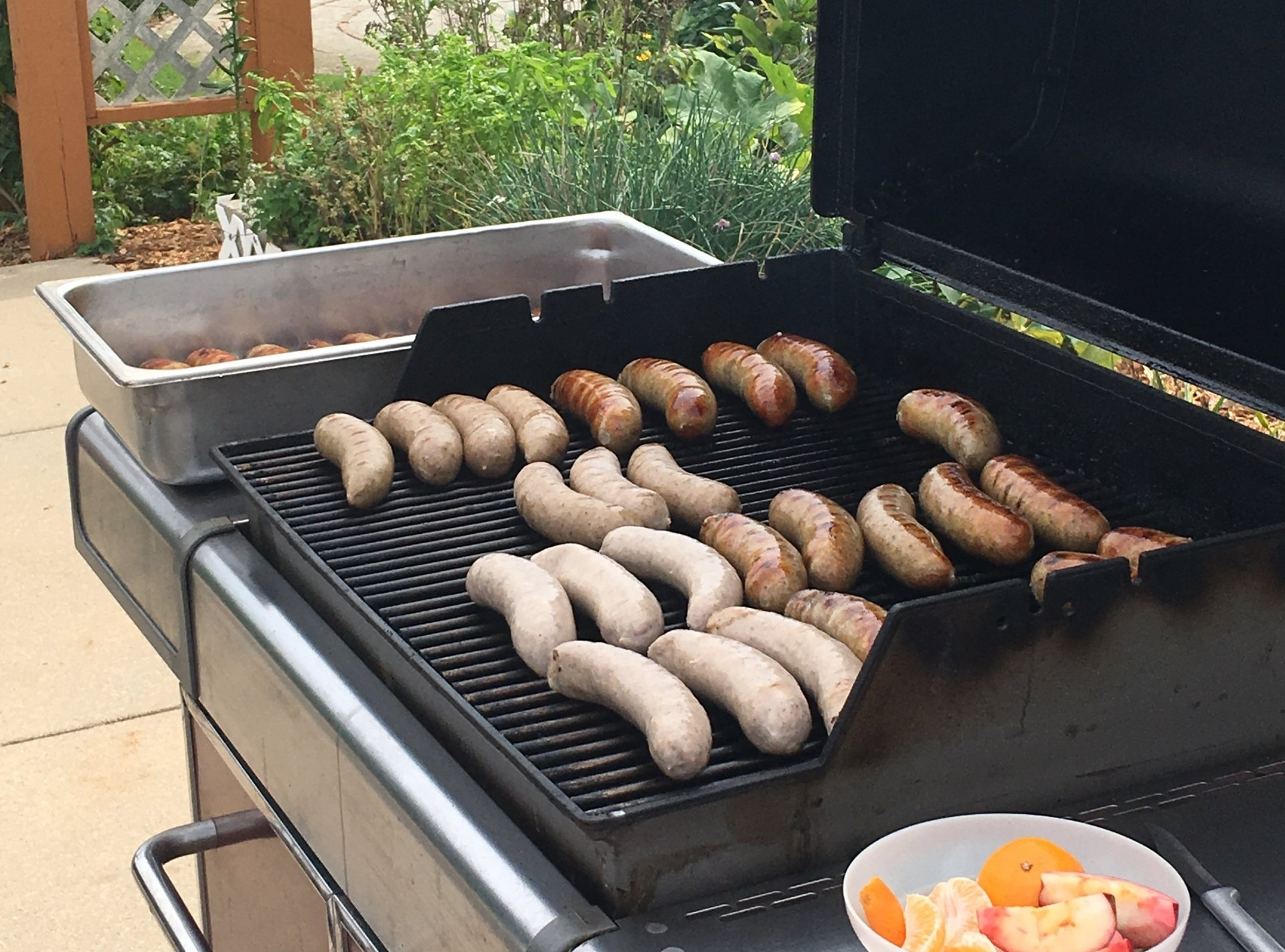 Grill with sausages cooking. Some already browned, others pale. A metal pan of sausages, and a bowl of fruit.