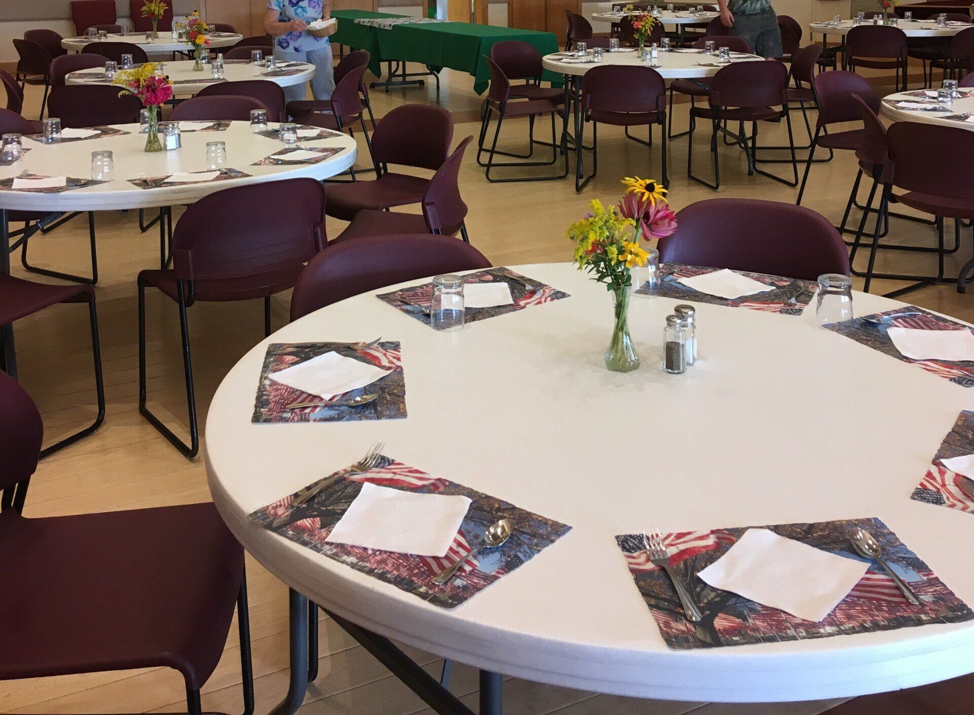 Round tables set with placemats, napkins, glasses, and flowers in a community hall.