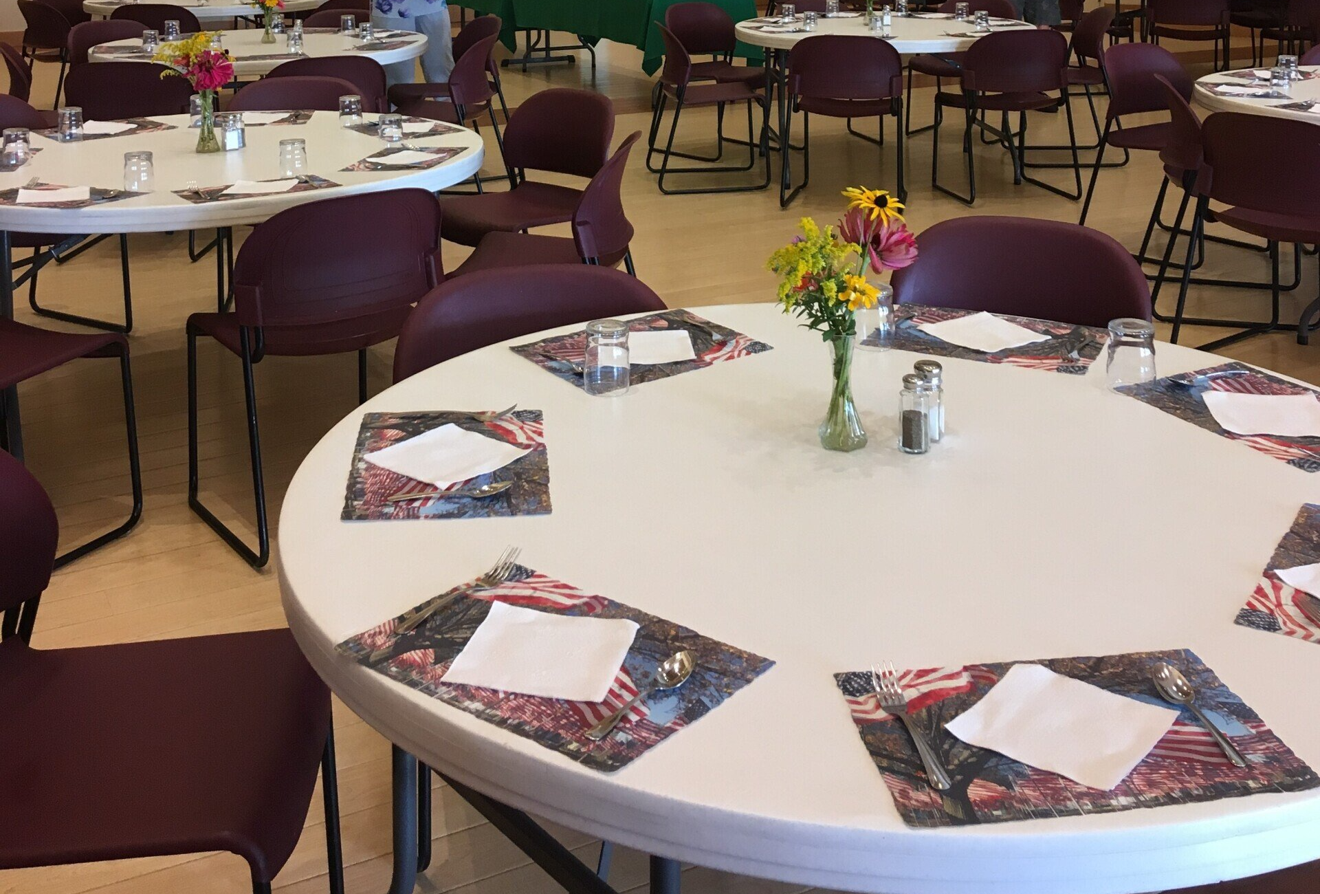 Round tables set for a meal with placemats, napkins, and flowers; chairs surround the tables in a dining hall.