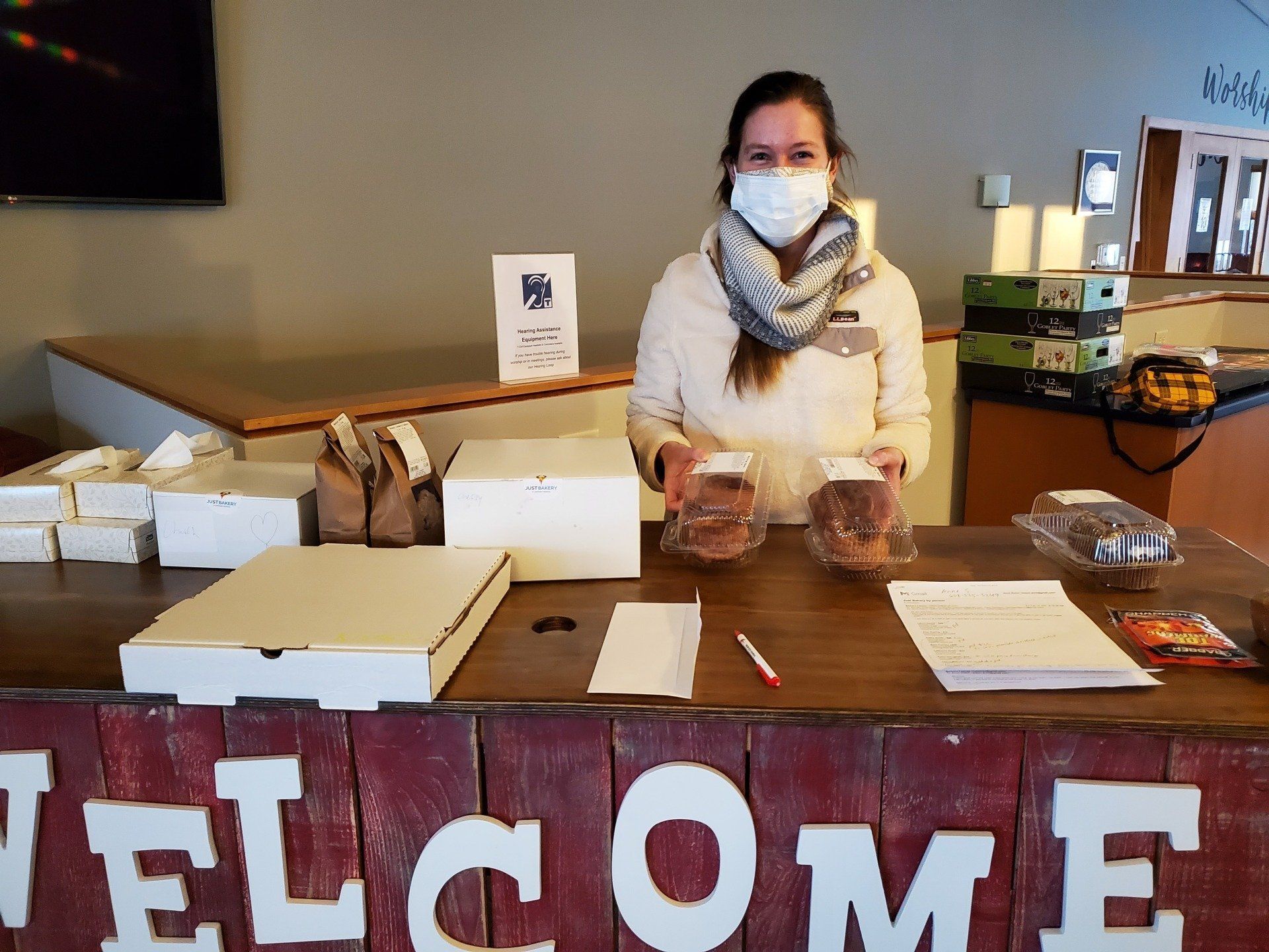 Woman in mask holds baked goods at a table with