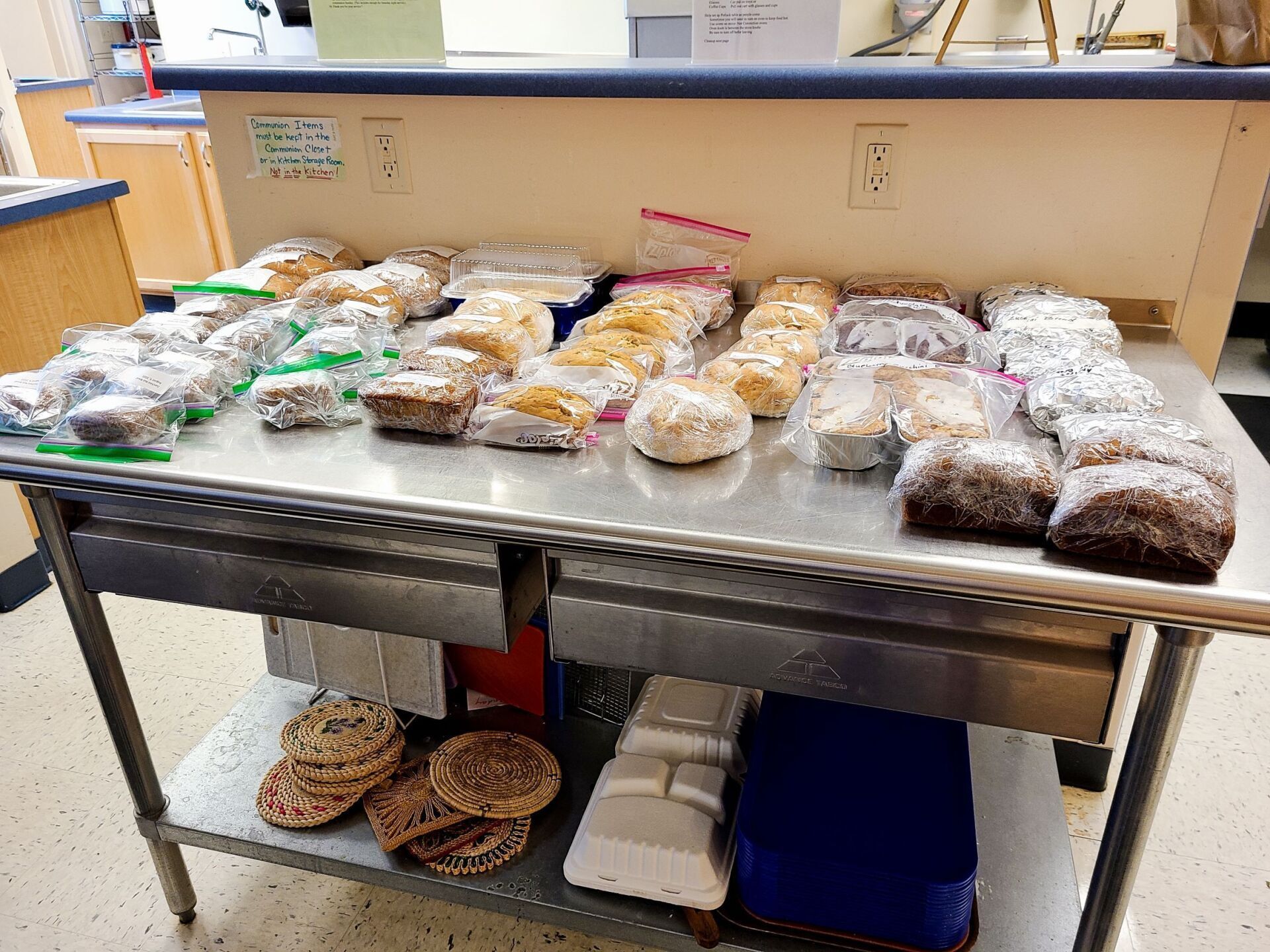 Baked goods on a metal table, wrapped in plastic. Some cookies below on a shelf.