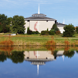 Modern church building reflected in calm water. Beige exterior with steeple and cross symbol.