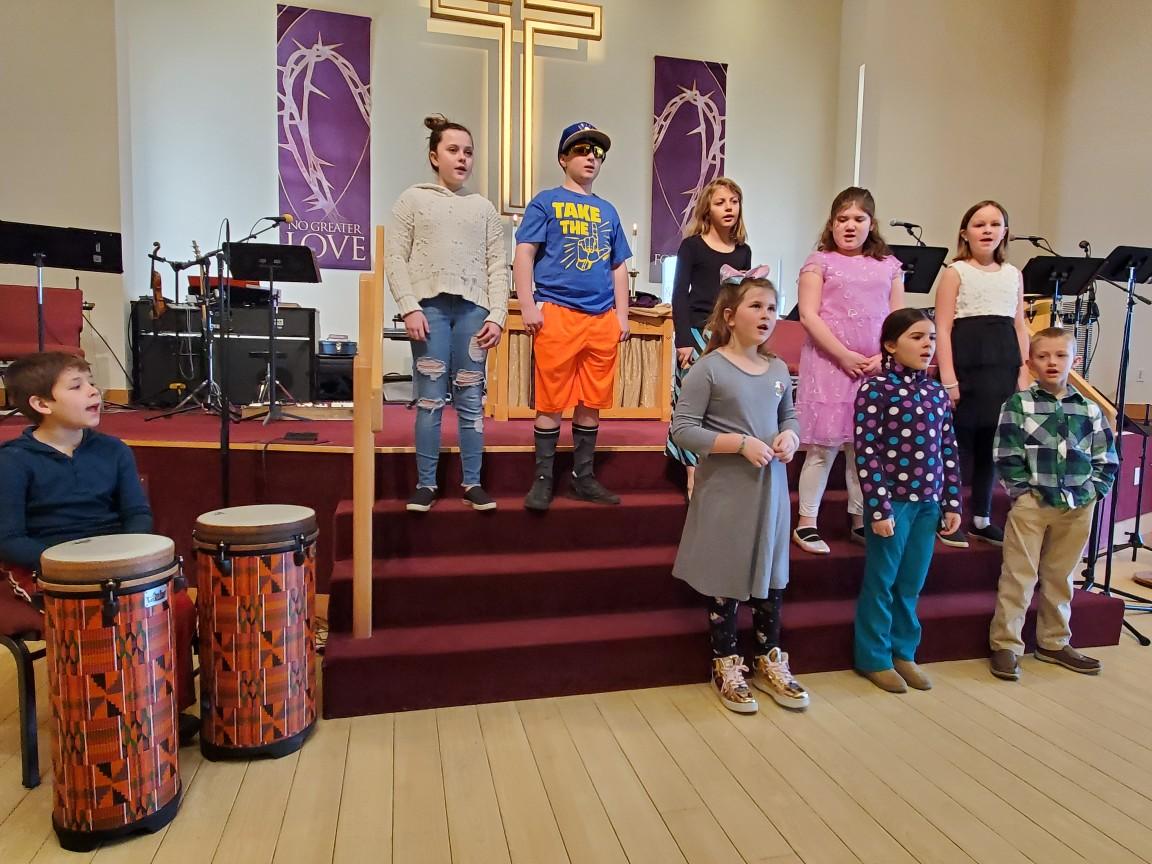 Children singing on a church stage with drums, instruments, and a cross backdrop.