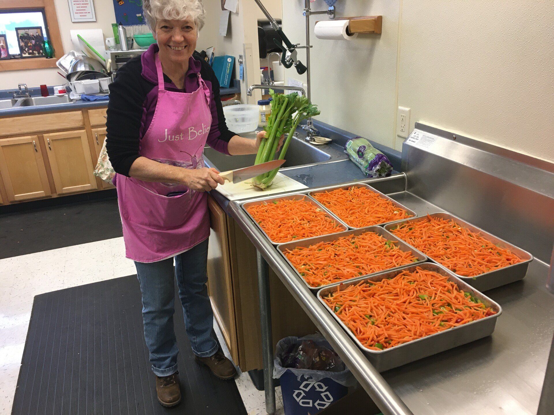 Woman in pink apron chopping celery in a kitchen. Six trays of shredded carrots are nearby.