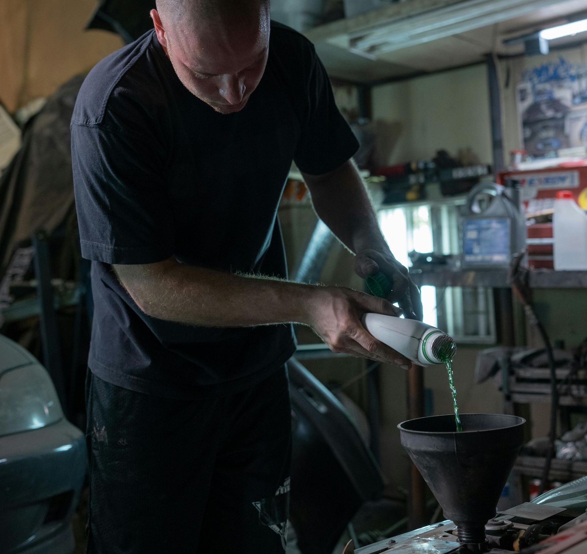 Man in a garage pouring green liquid from a bottle into a car's engine using a funnel.