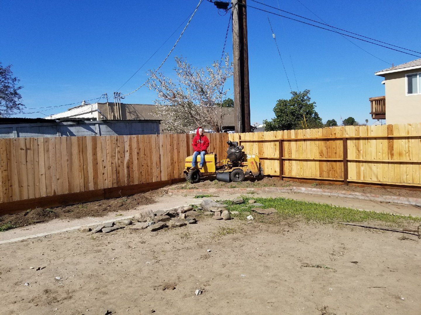 Person in red operating a mower in a fenced dirt yard under a clear blue sky