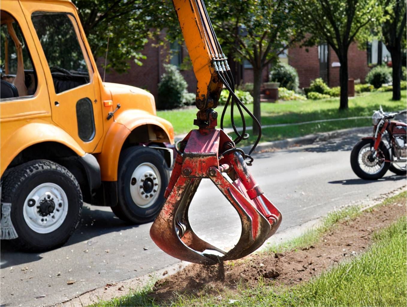 Orange crane lifting a red excavator bucket from a roadside trench in a suburban street
