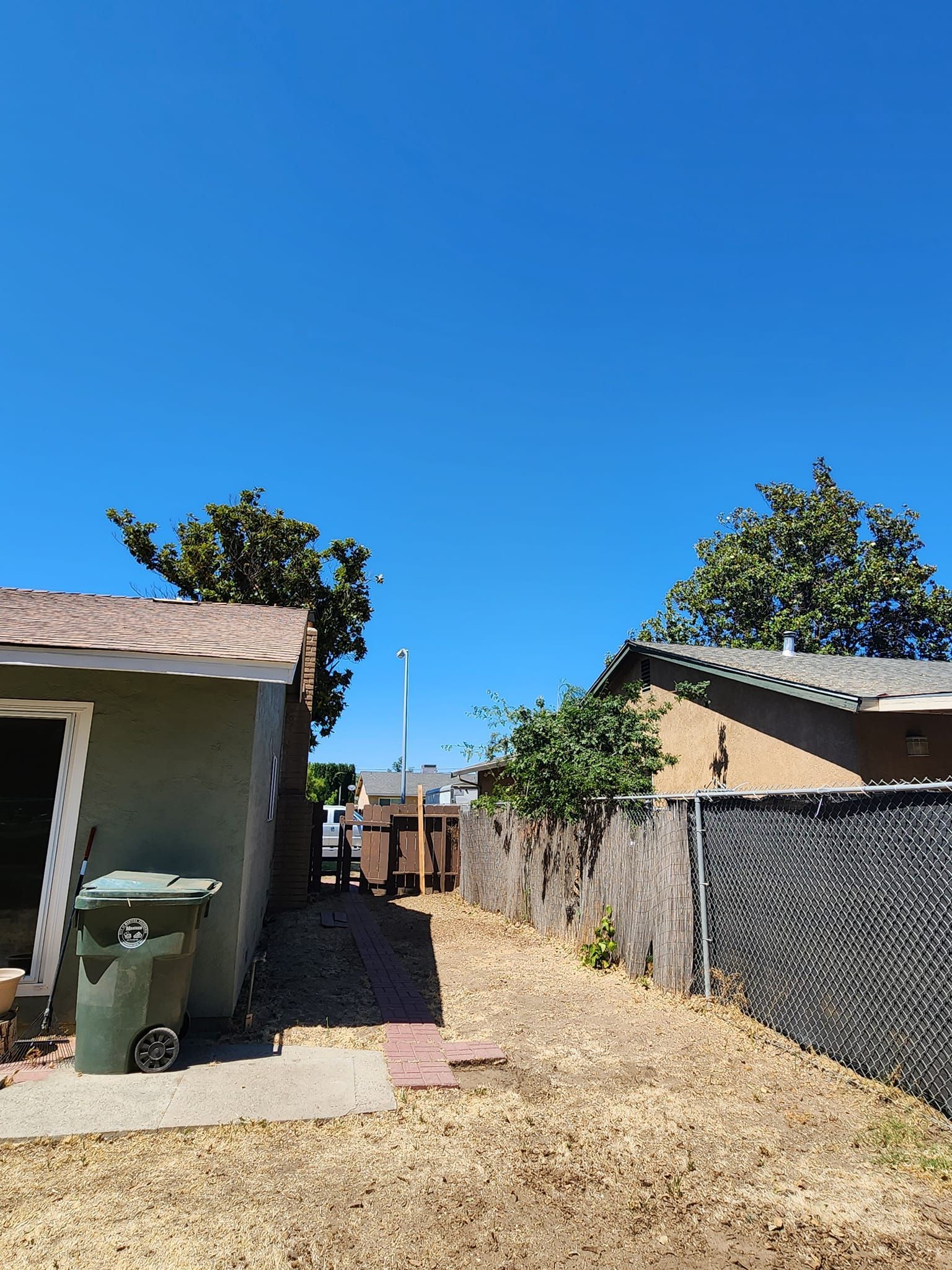 Narrow backyard alley with gravel, two houses, a trash bin, and a bright blue sky.