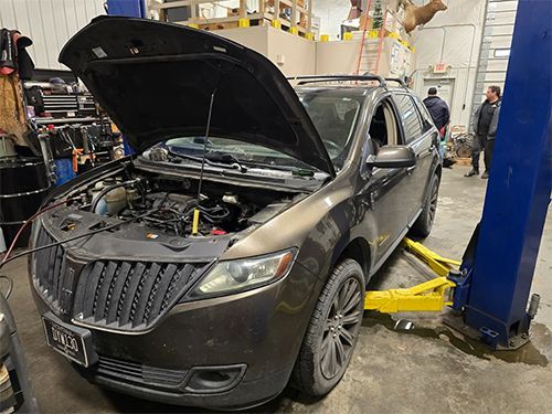 Dark SUV with hood open in an auto repair shop, lifted by a yellow jack under a blue hoist.
