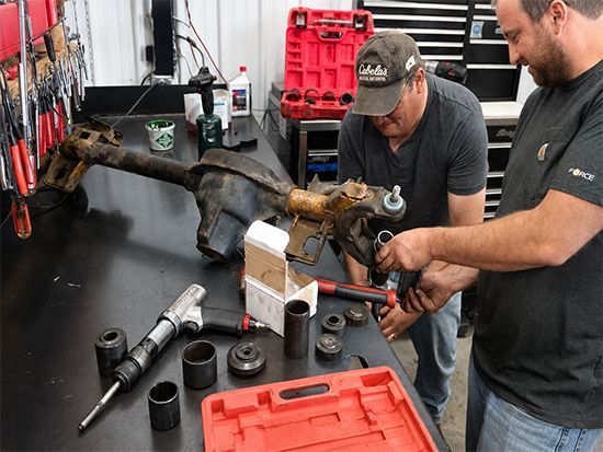 Two mechanics working on a car axle in a workshop, with tools and red parts trays on a bench.