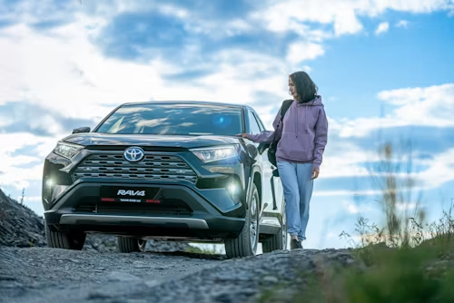 SUV on a rocky road with a person in a purple jacket standing beside it under a blue sky