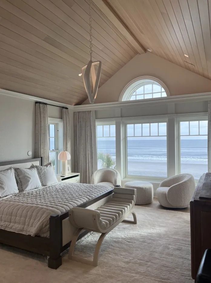 Bright beach-view bedroom with vaulted ceiling, large windows, and white furniture