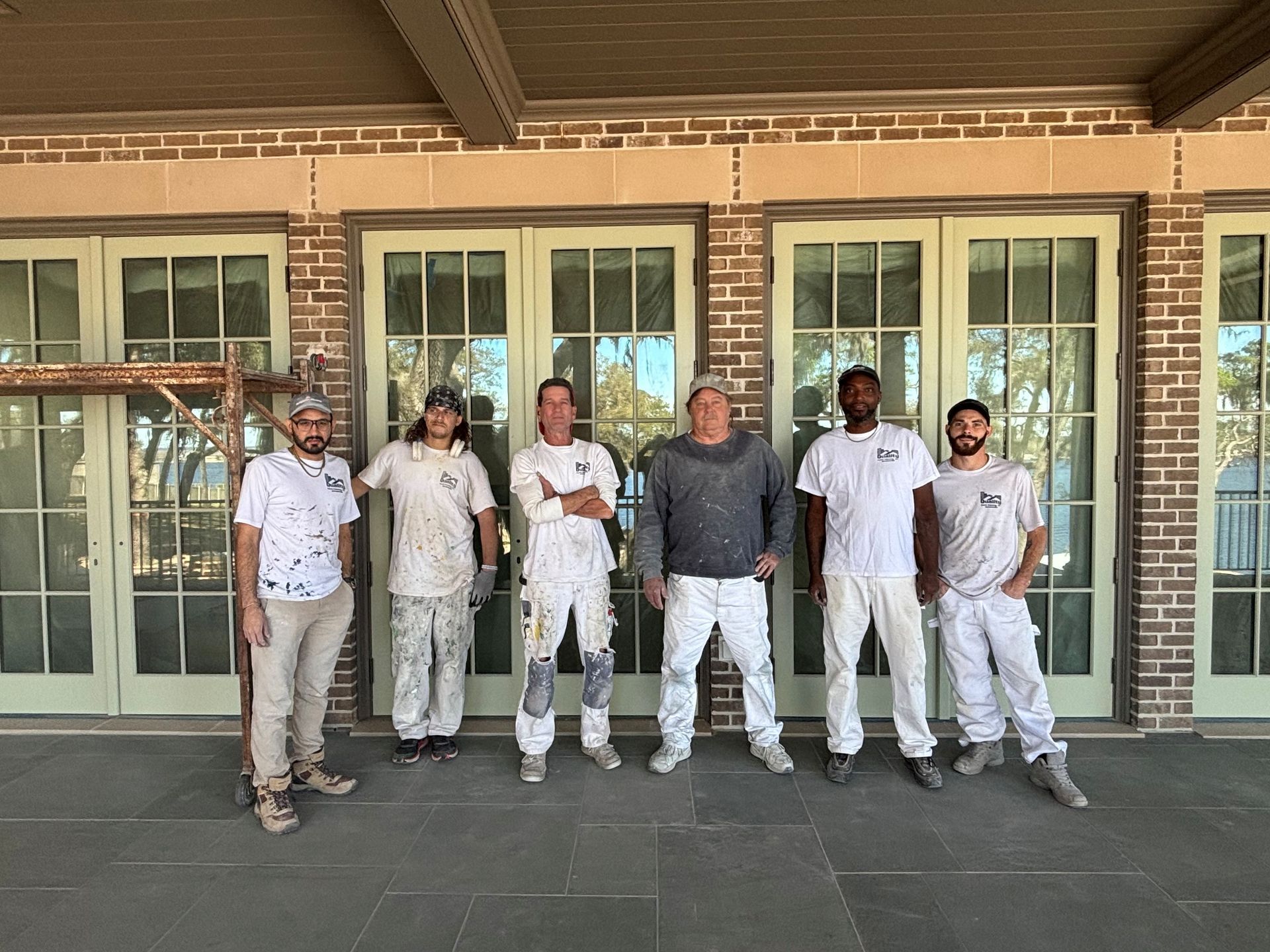 Group of painters in paint-splattered white outfits, standing in front of glass doors and a brick facade.